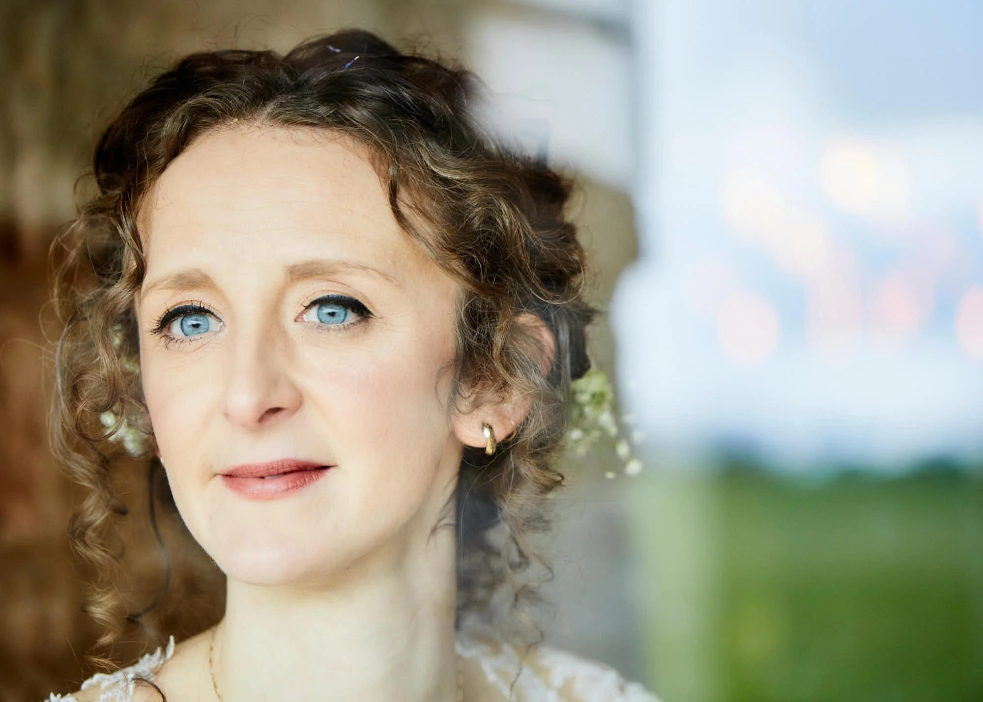 A close-up of a woman with curly, shoulder-length brown hair, blue eyes, light makeup, wearing small earrings and a white lace top, outdoors with a blurred background of nature.