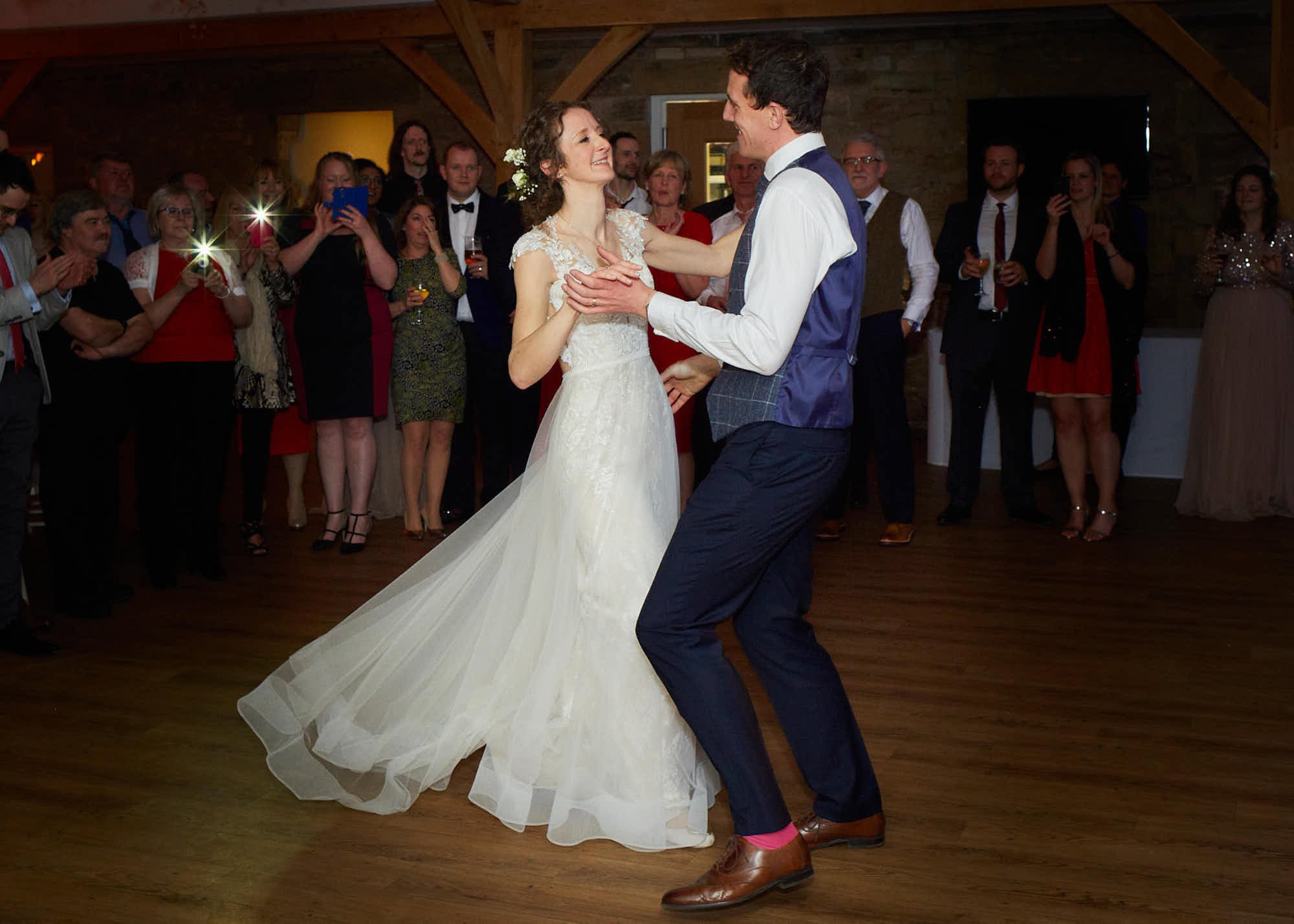 A bride and groom dancing at their wedding reception with guests watching and taking photos, inside a rustic venue.