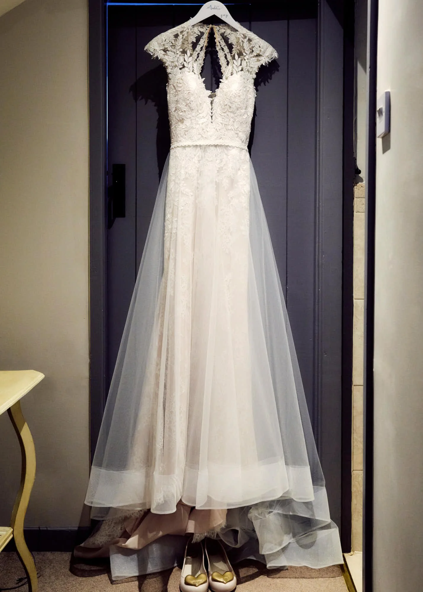 A wedding dress hanging on a hanger in front of a dark background, with beige high-heeled shoes with gold heart-shaped decorations at the toe positioned underneath.