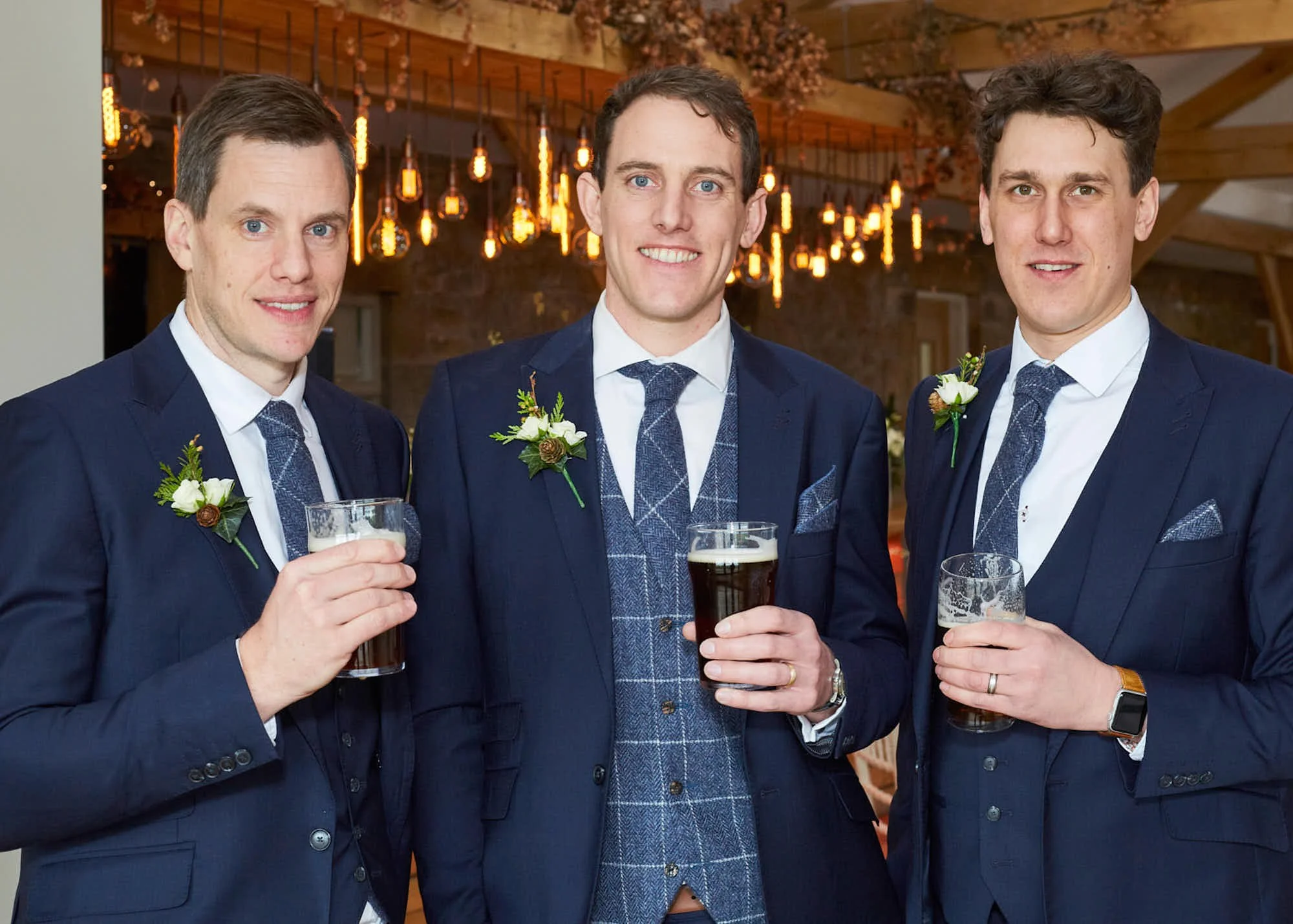 Three men in suits and ties at a social event, holding glasses of beer, smiling, with a warm, decorated background featuring hanging lights and wooden accents.