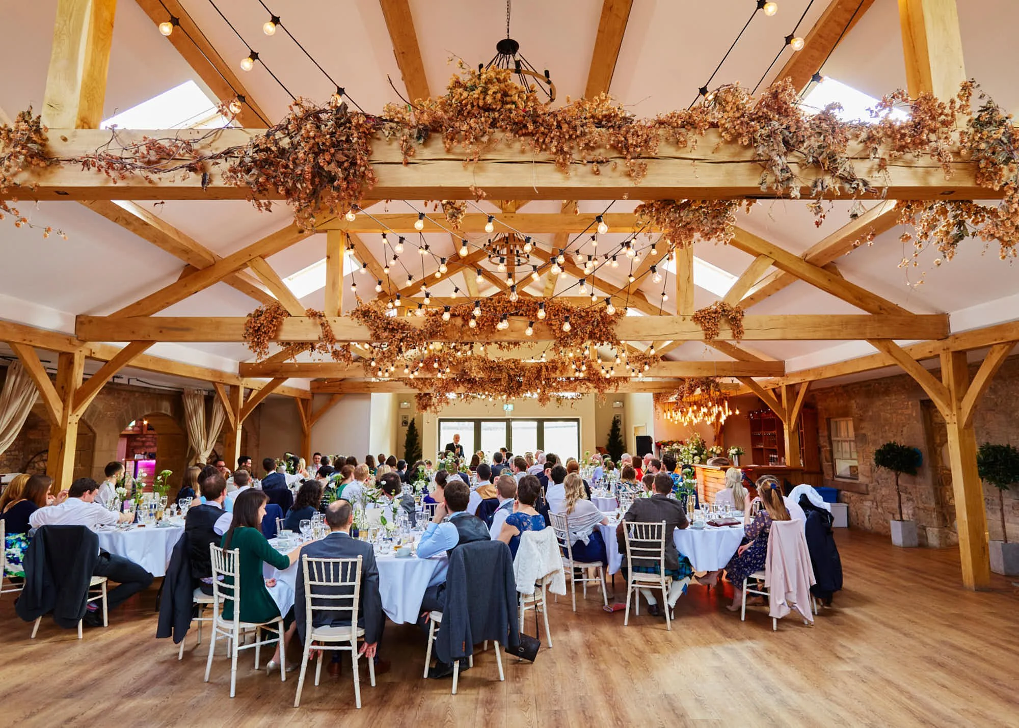 A large banquet hall filled with many round tables set for a formal event with white tablecloths, plates, glasses, silverware, and floral centerpieces. Guests are seated, dressed in formal attire, listening to a speaker at a podium. The ceiling has w