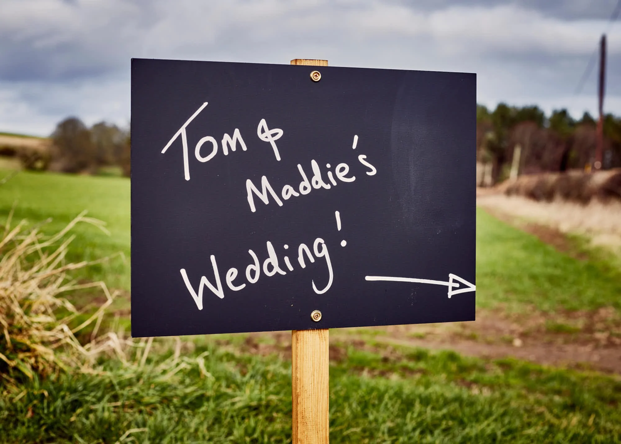 A sign on a wooden post in a rural area reading 'Tomp & Maddie's Wedding' with an arrow pointing to the right, with cloudy skies in the background.