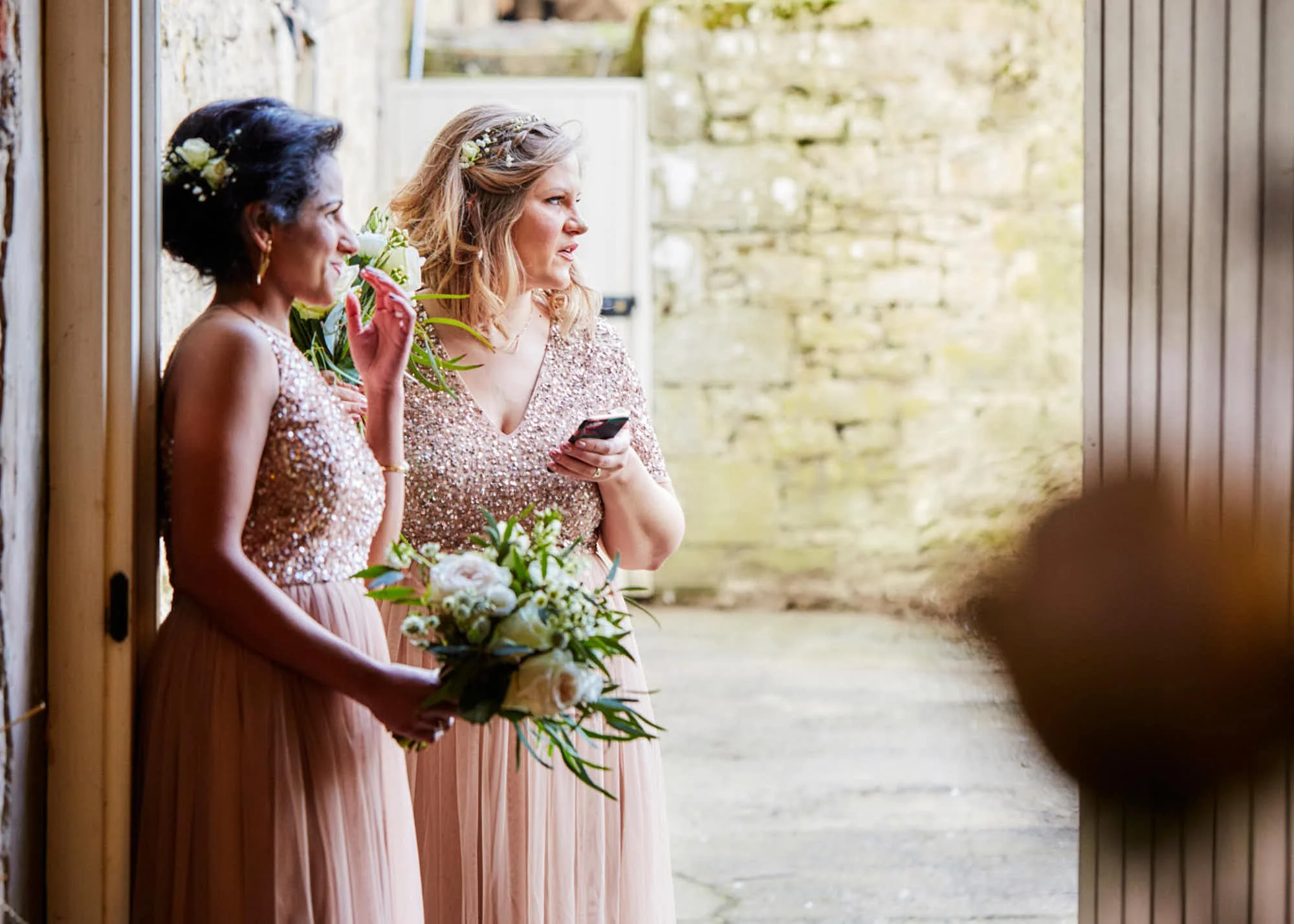 Two women dressed in pink, sequined dresses, standing by a wooden wall, holding bouquets of flowers, with one using her phone, at what appears to be a wedding or special event.