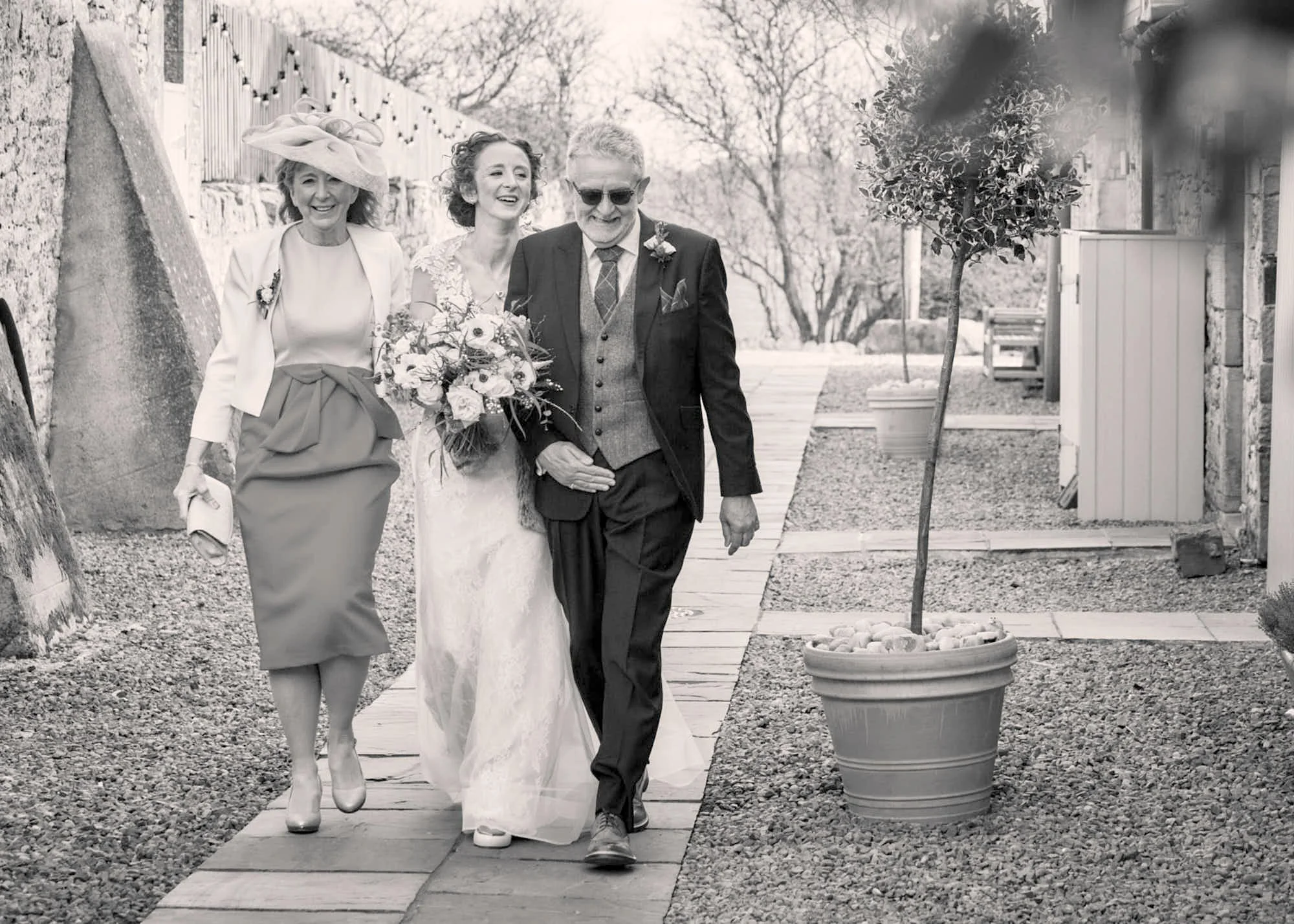 A black and white photo of three people walking outdoors on a pathway. The group includes a woman in a hat and dress, a bride in a wedding gown holding a bouquet, and a man in a suit with sunglasses. They are smiling and appear happy.