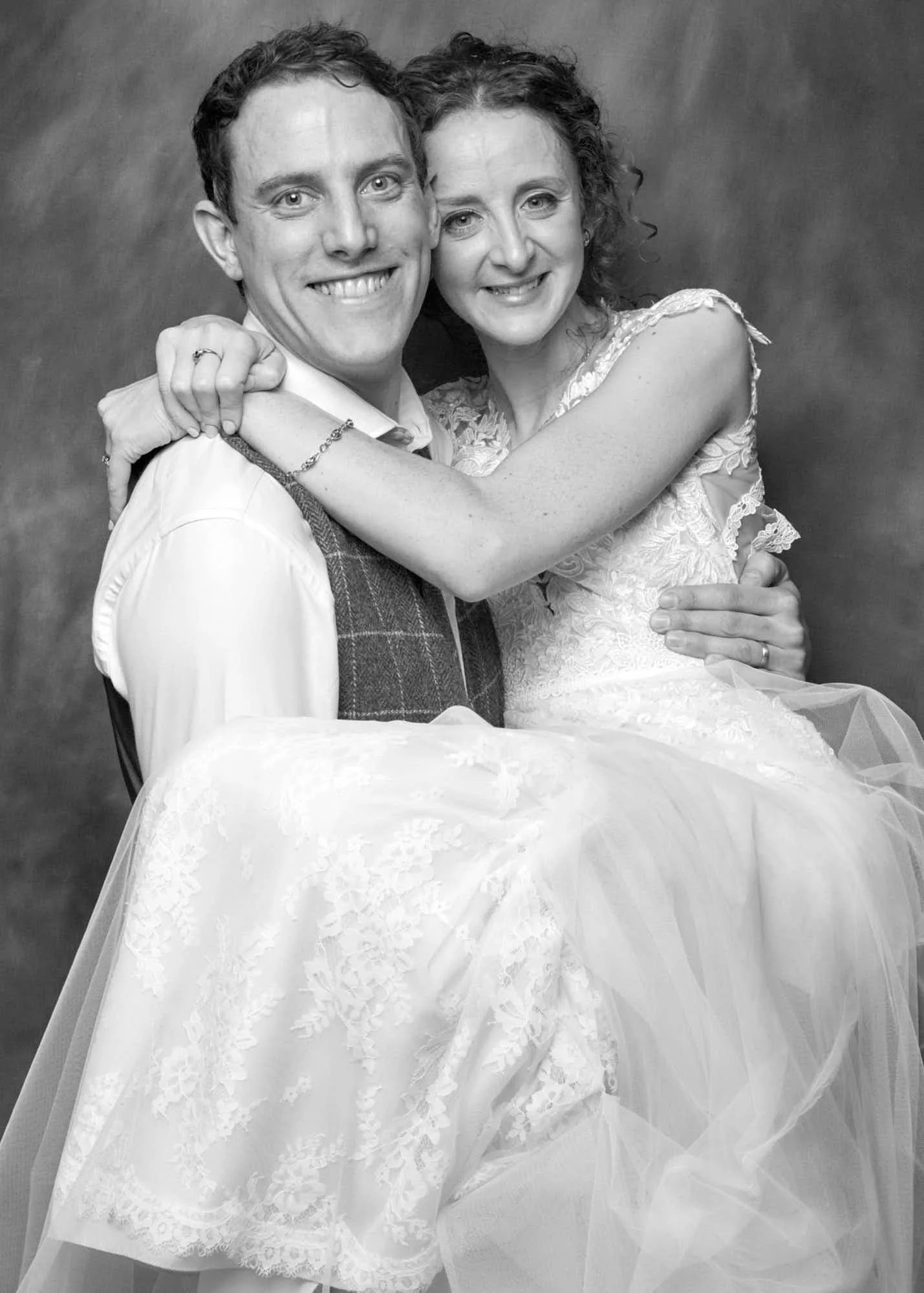 Black and white photo of a smiling young man and woman embracing, the woman is wearing a wedding dress, and the man is in a vest and shirt, against a plain background.