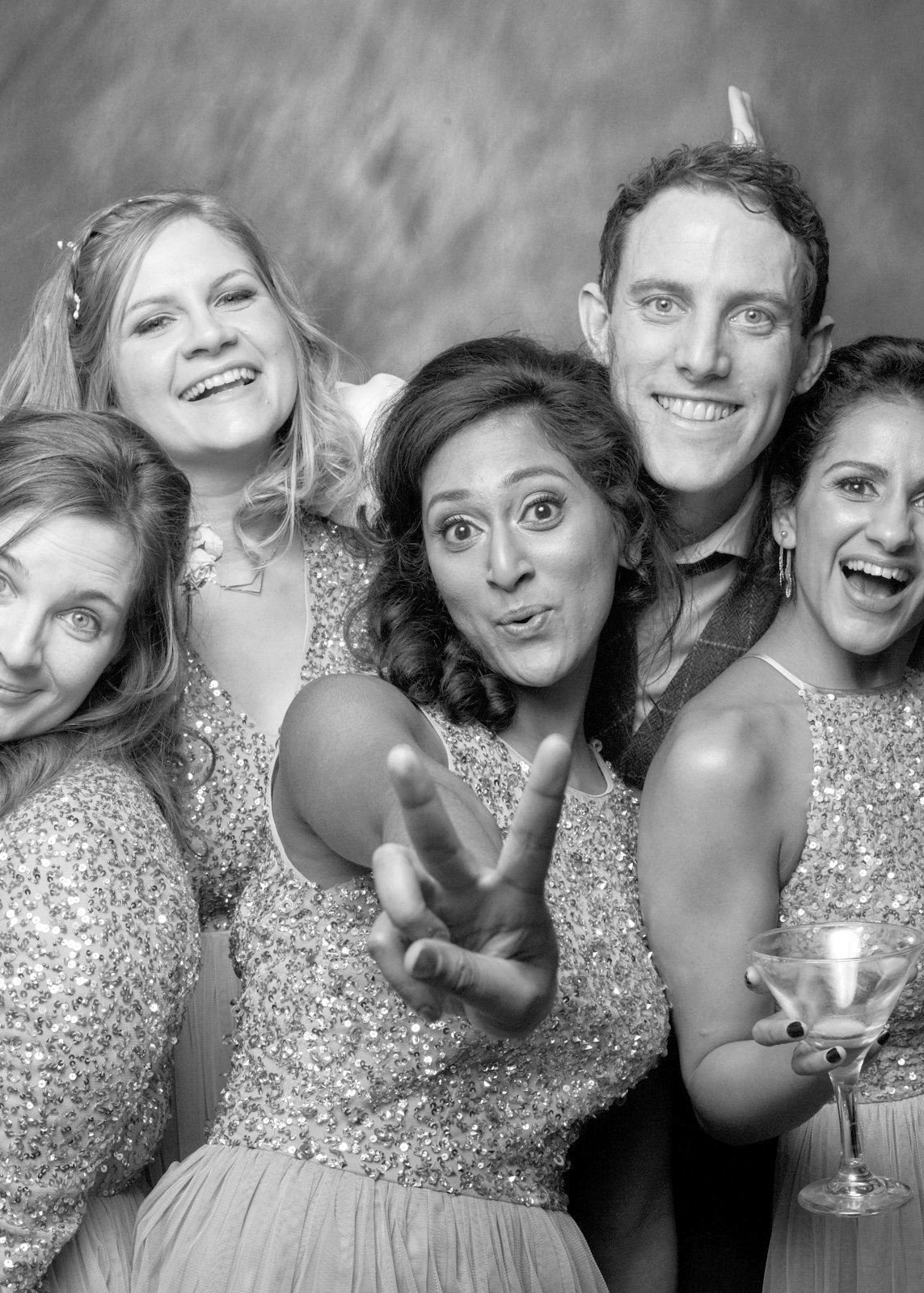 Group of five women and one man at a celebration, smiling and laughing, with one woman making a peace sign and holding a cocktail glass, dressed in sparkly outfits.