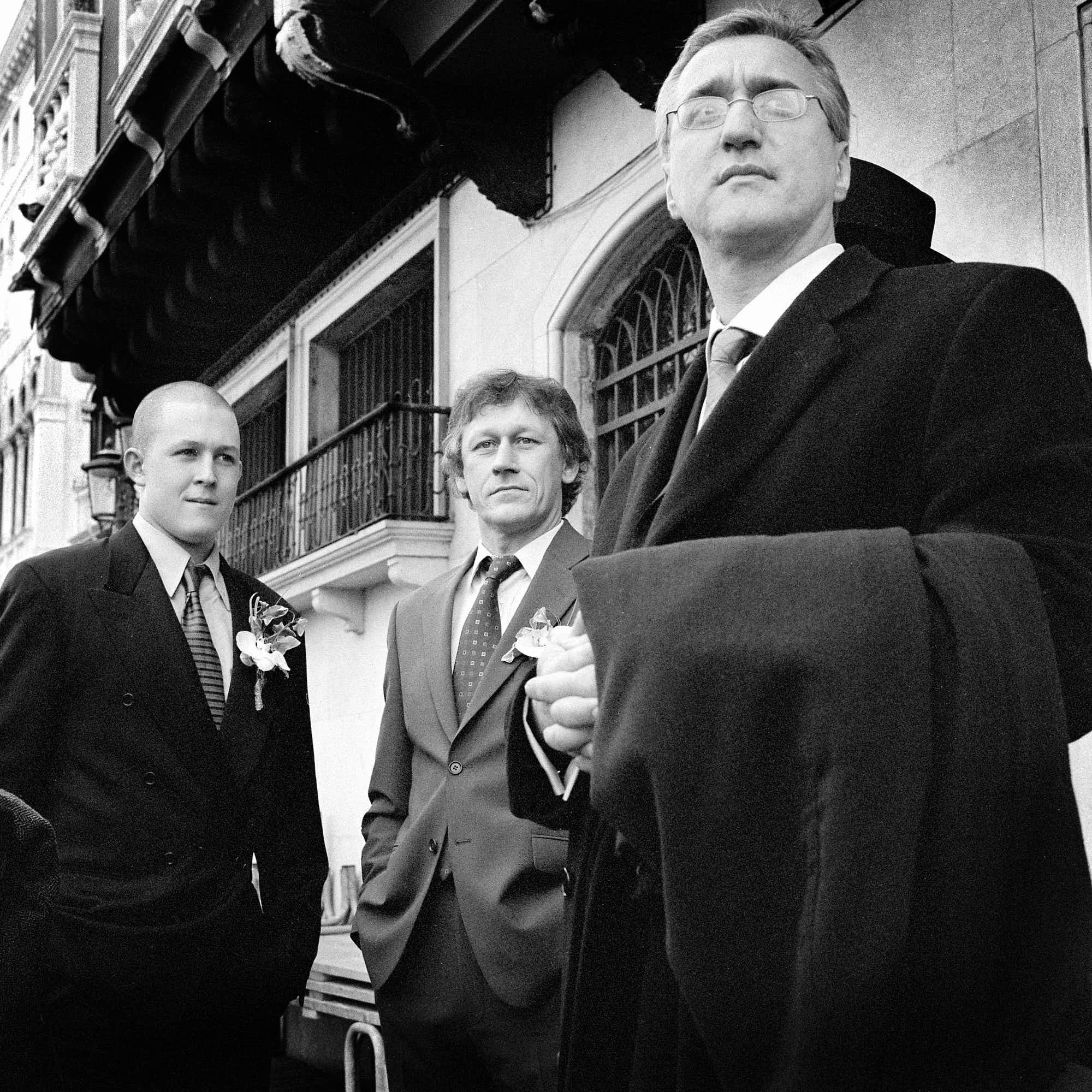 Black and white photo of three men standing outdoors near a building with balconies. The man in the foreground is wearing glasses and a suit with a tie, looking off into the distance with a serious expression. The second man, slightly behind, has cur