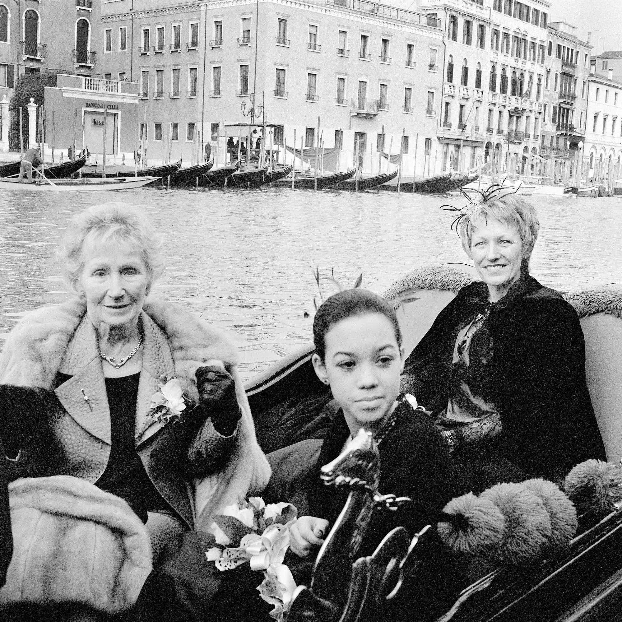 Black and white photo of several gondolas docked along a canal in Venice, Italy, with historic buildings and a small pier in the background, and a woman and children in the foreground.