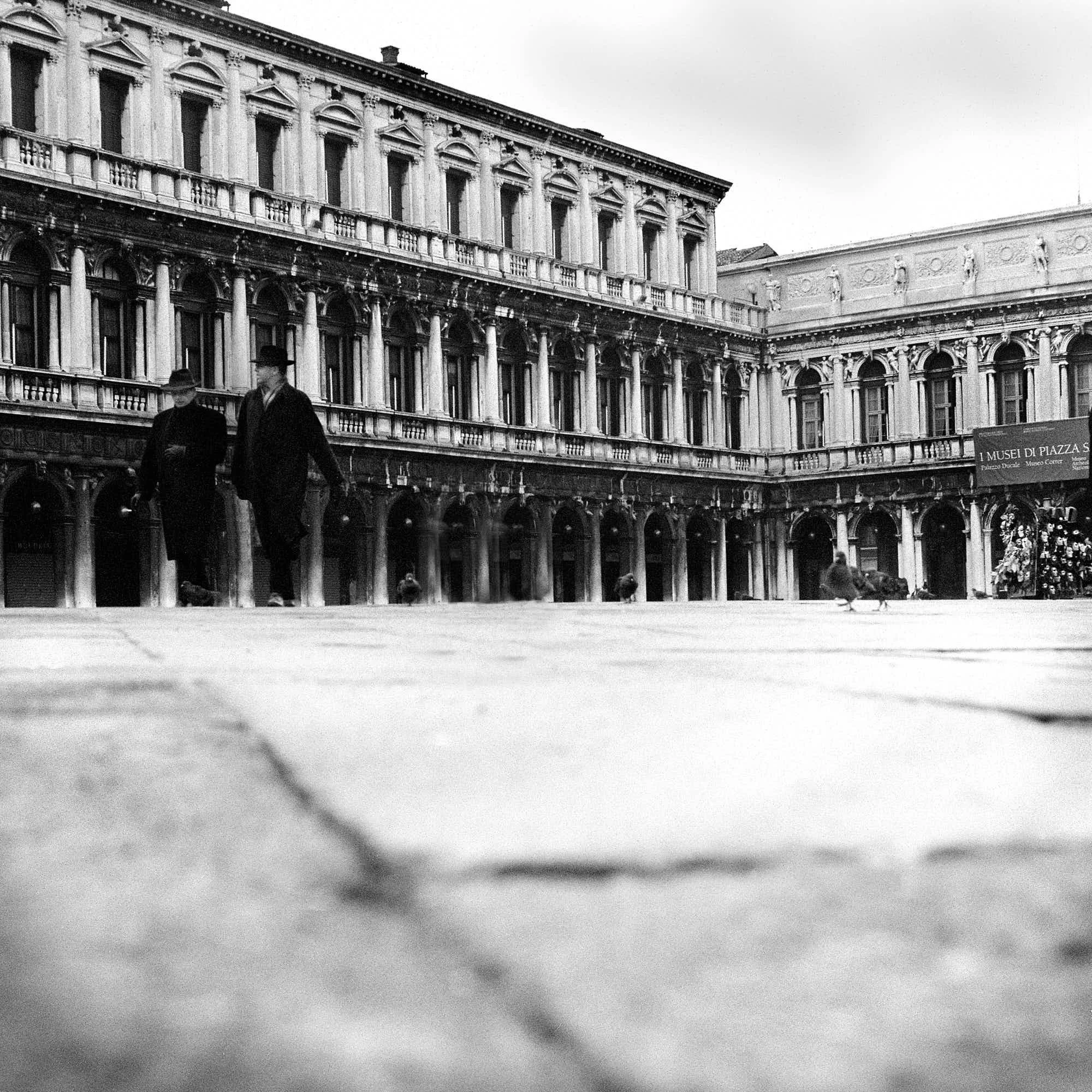Black and white photograph of an ornate, historic building with multiple arched windows and decorative columns. Two men wearing coats and hats walk in the foreground. There is a sign indicating the location of the museums at the bottom right.