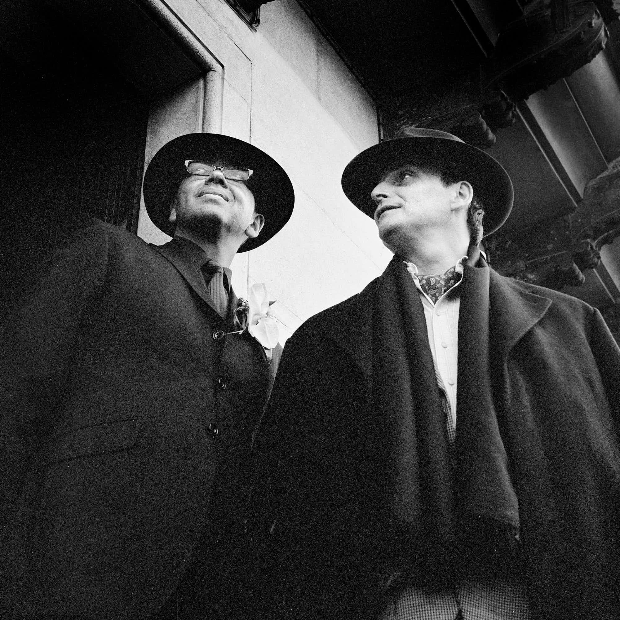 Two men in formal attire with black hats standing and talking on a city street.