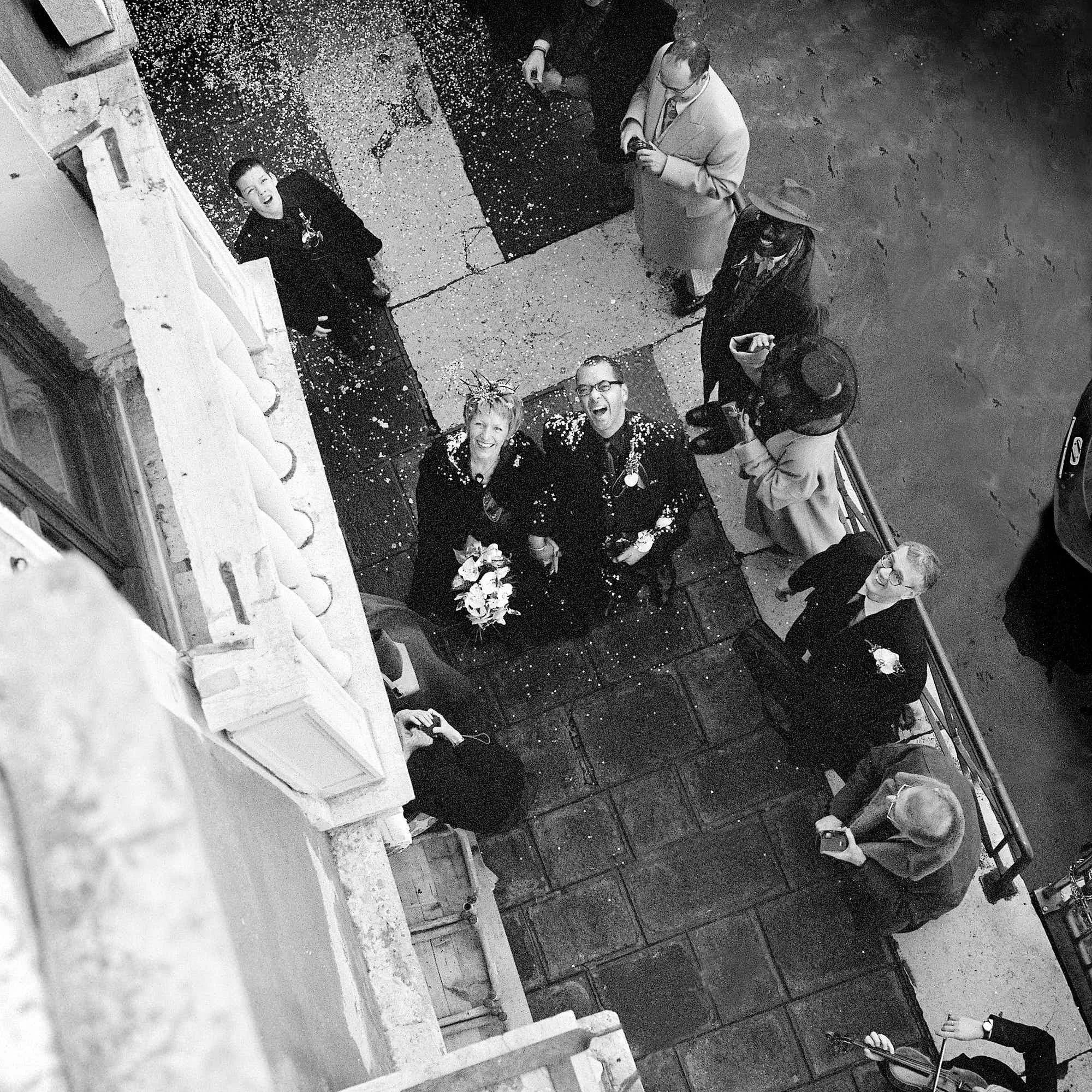 A black and white photo taken from a high angle showing five people outdoors at night, standing on a gravel and concrete surface, with a building wall visible on one side. The group appears to be celebrating, with some smiling or laughing, and confet