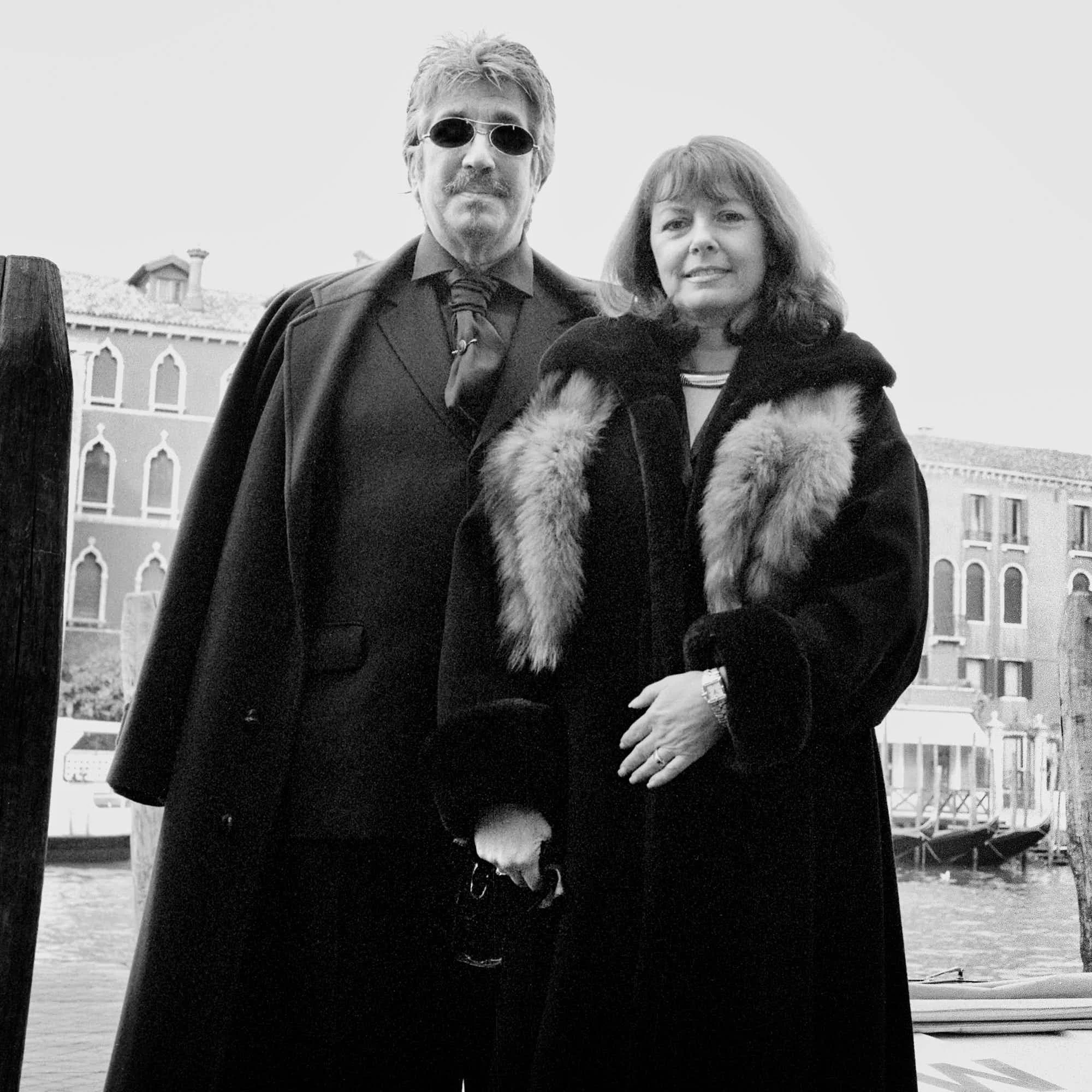 A black and white photo of a man with sunglasses and a woman with wavy hair, standing outdoors with buildings in the background.