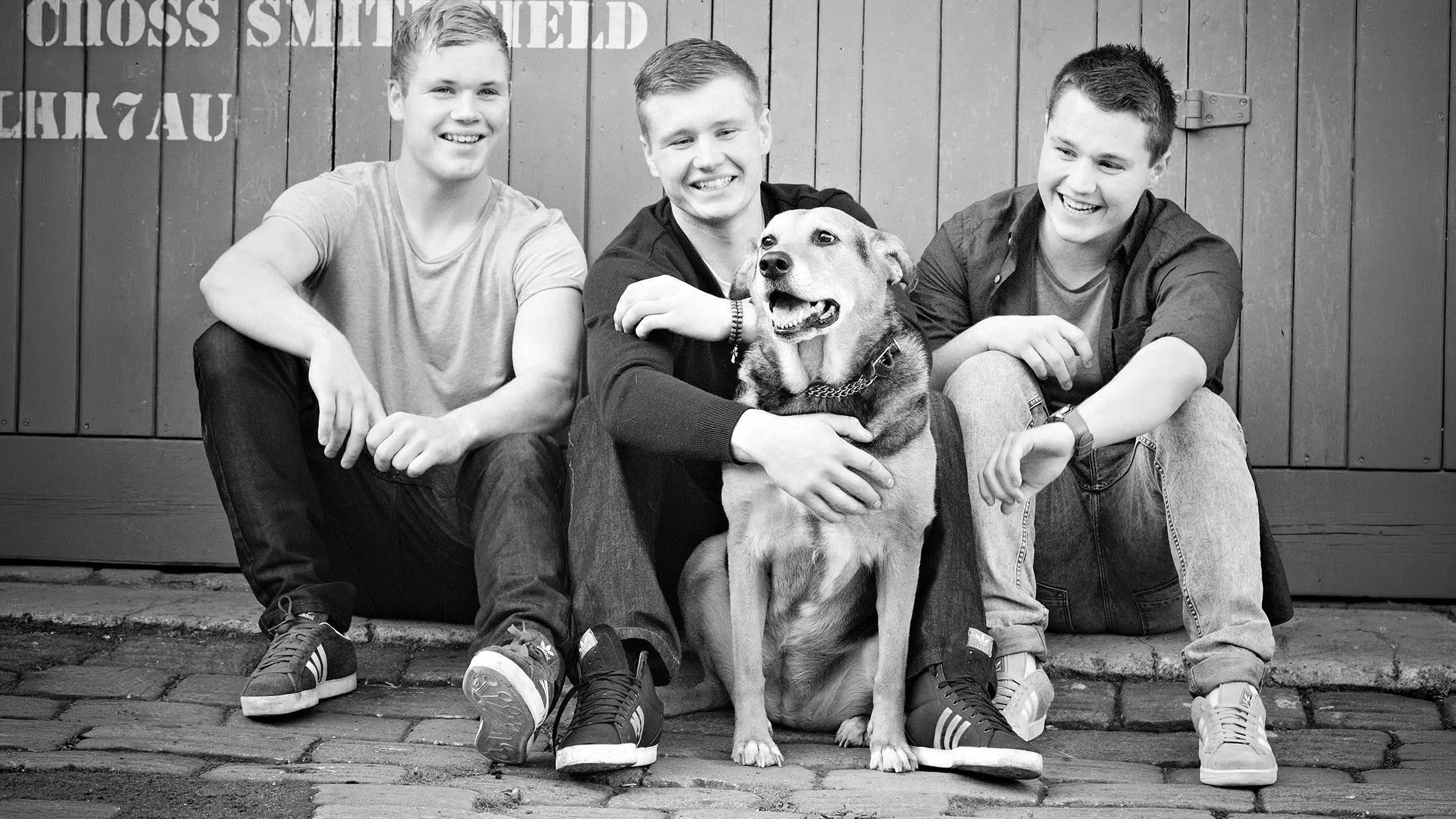 Four young men sitting on a curb with a dog in front of a wooden wall, smiling and looking at the camera, in black and white.