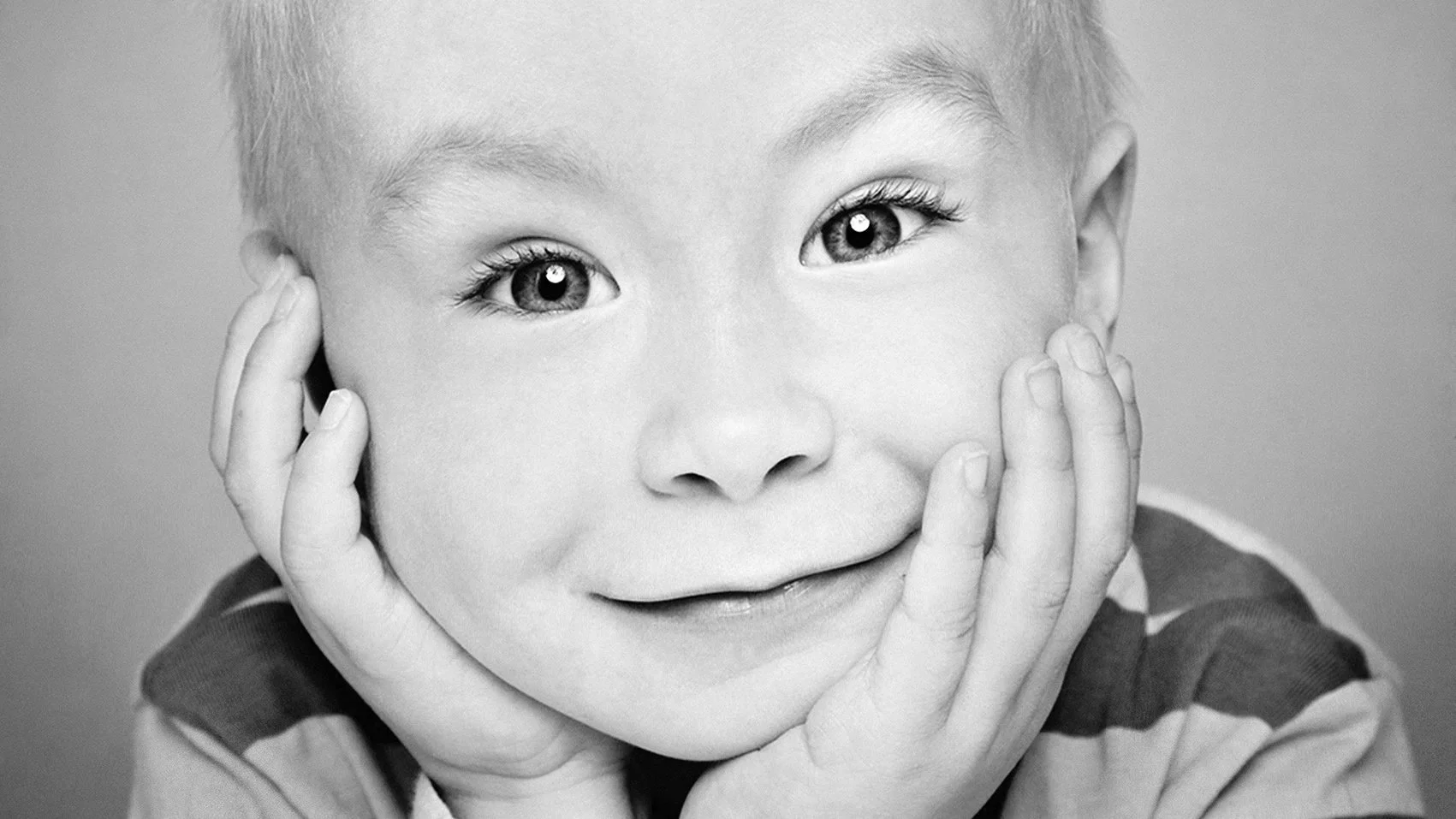 A close-up black and white photo of a smiling child with hands on cheeks, showing expressive eyes and a joyful expression.