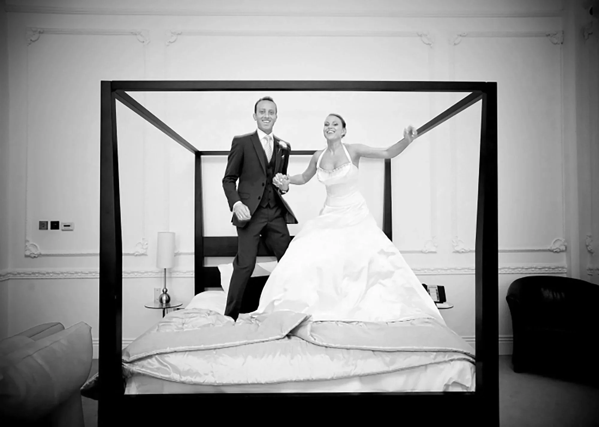 Black and white photo of a smiling man in a suit and woman in a wedding dress holding hands on a bed in a bedroom with ornate wall decor.