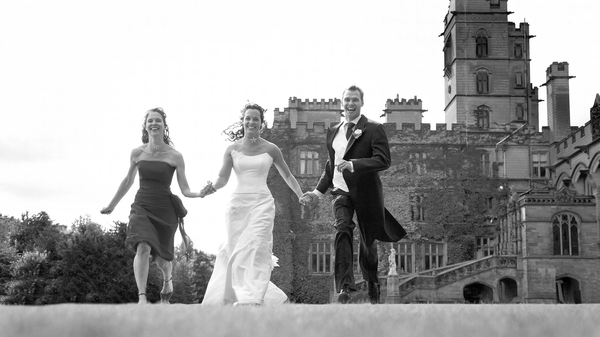 A joyful bride and groom holding hands, walking with two women, all smiling, outside a historic castle or mansion in black and white.