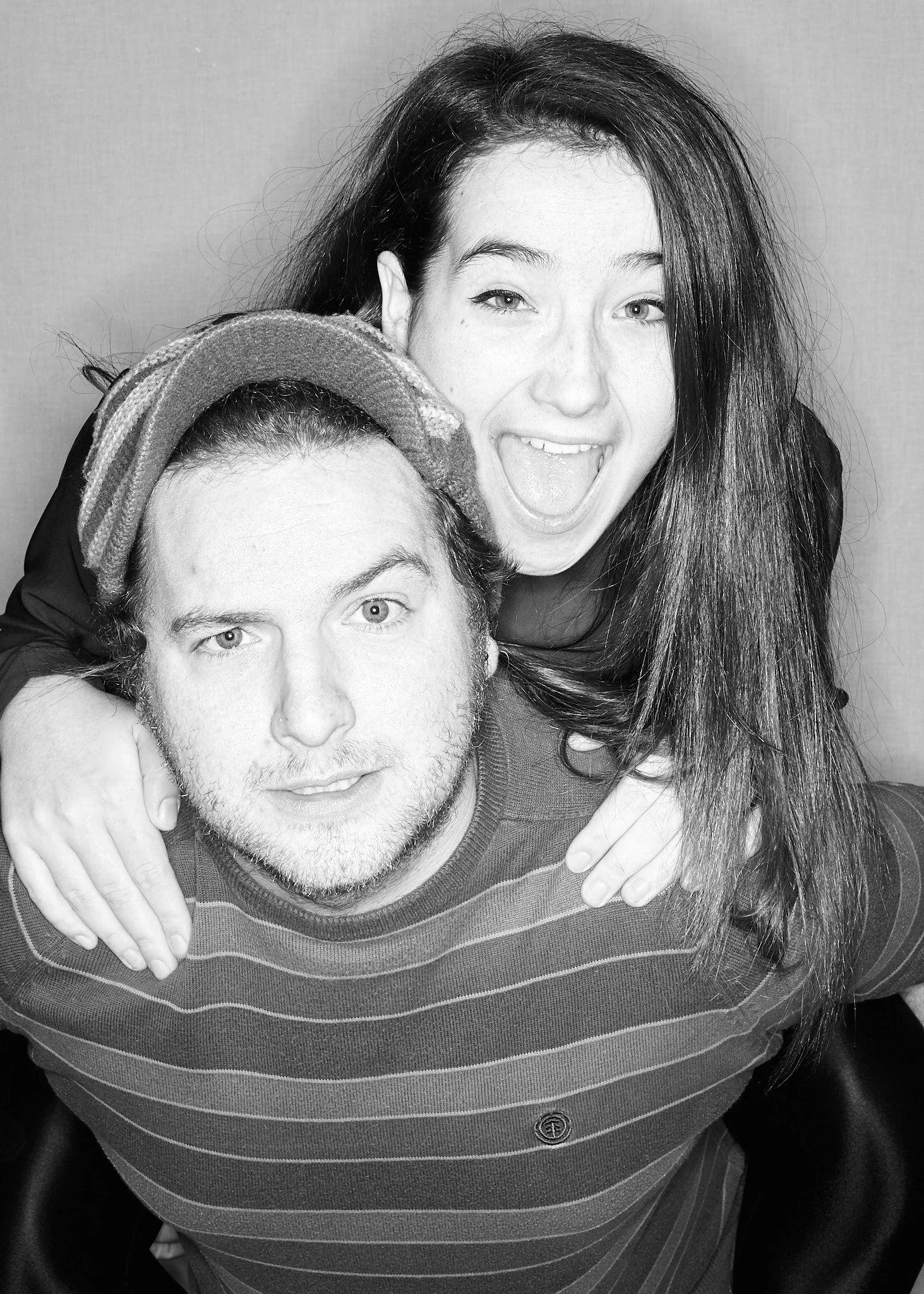 Black and white photo of a young woman with long dark hair, smiling with her mouth open and tongue out, hugging a young man with light-colored hair and light eyes, wearing a striped shirt, with a neutral background.