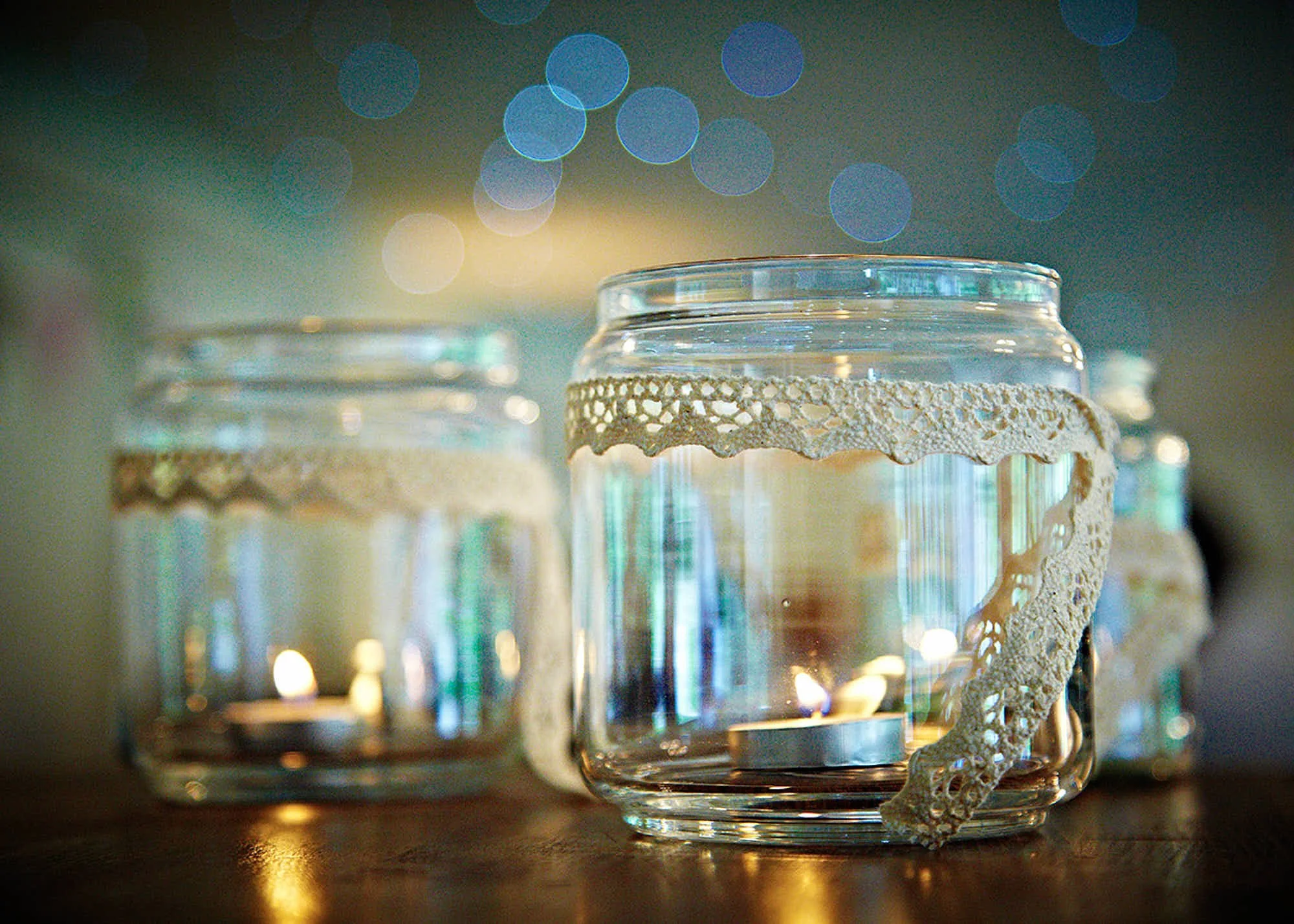 Close-up of three glass candles holders with lace decorations, lit candles inside, placed on a wooden surface with a blurred background of bokeh lights.