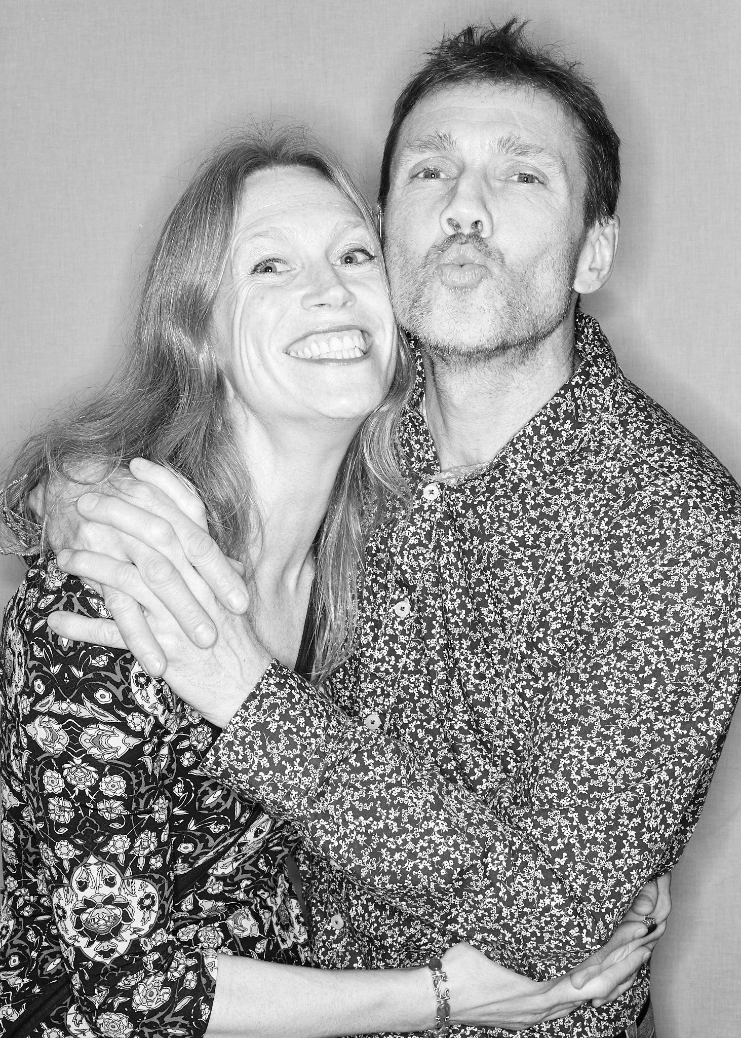 A black and white photo of a man and woman hugging and making playful faces at the camera, both wearing patterned shirts.