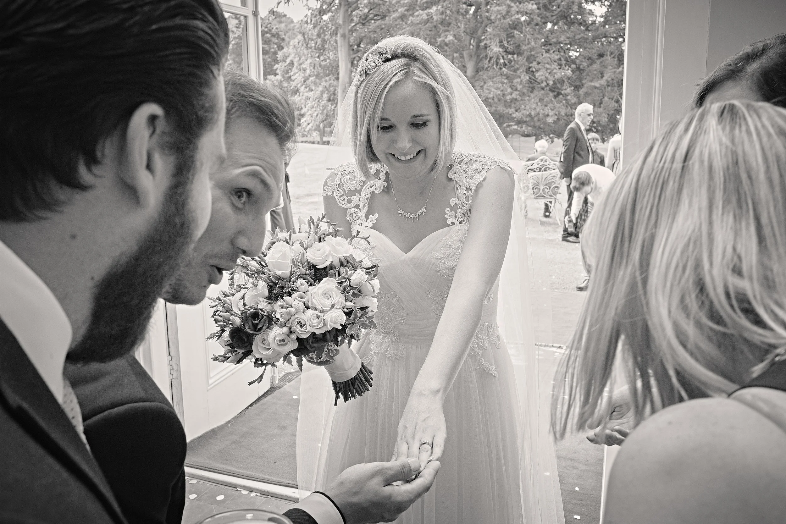 A bride on her wedding day, smiling as she receives a wedding ring from a man, with three women nearby, and a lush outdoor garden visible through the window.