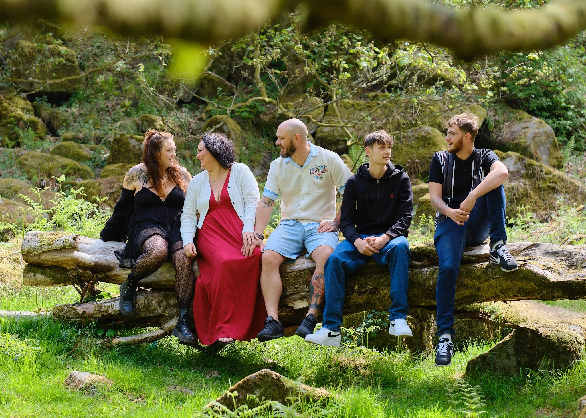 Six young people sitting on a fallen log in a forest, smiling and talking to each other.