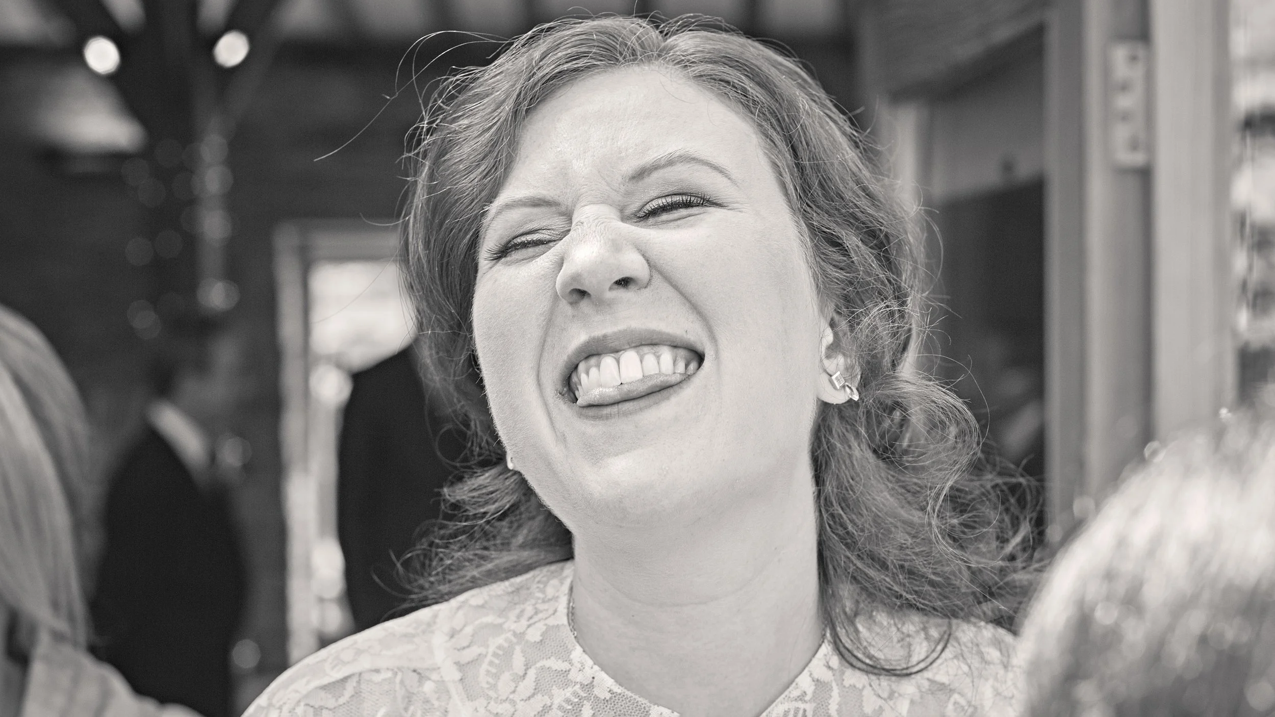 A woman smiling with her tongue out, wearing earrings and a lace top, in black and white.
