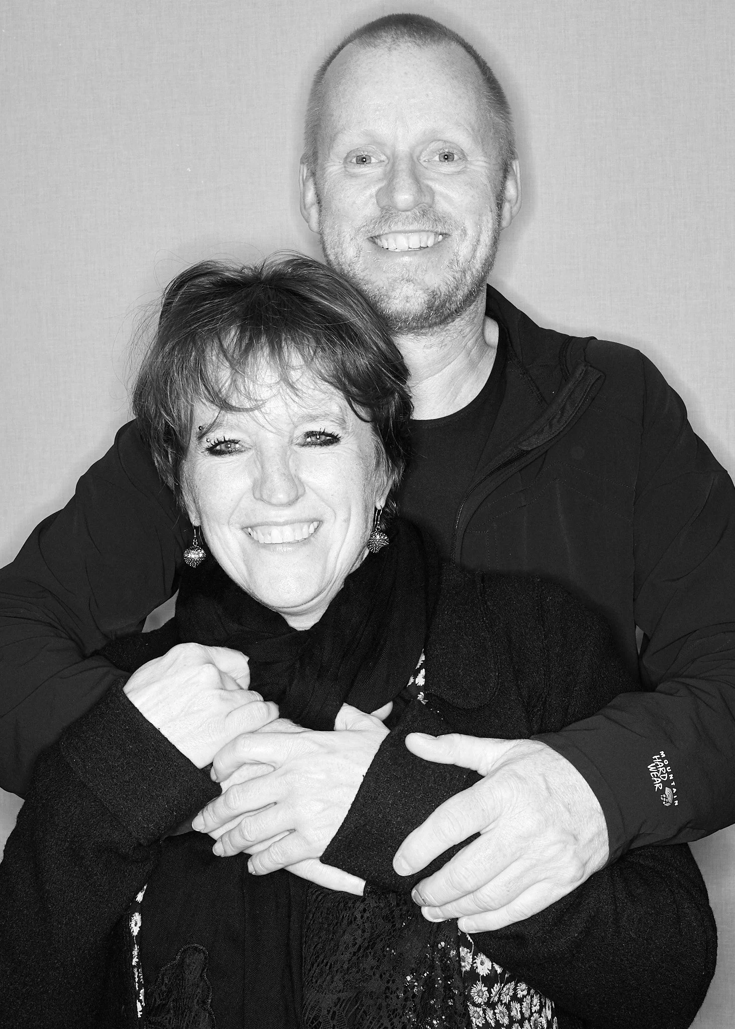 A black and white photo of a smiling man and woman hugging each other closely, with the woman's head resting on the man's shoulder. They are indoors against a plain wall.