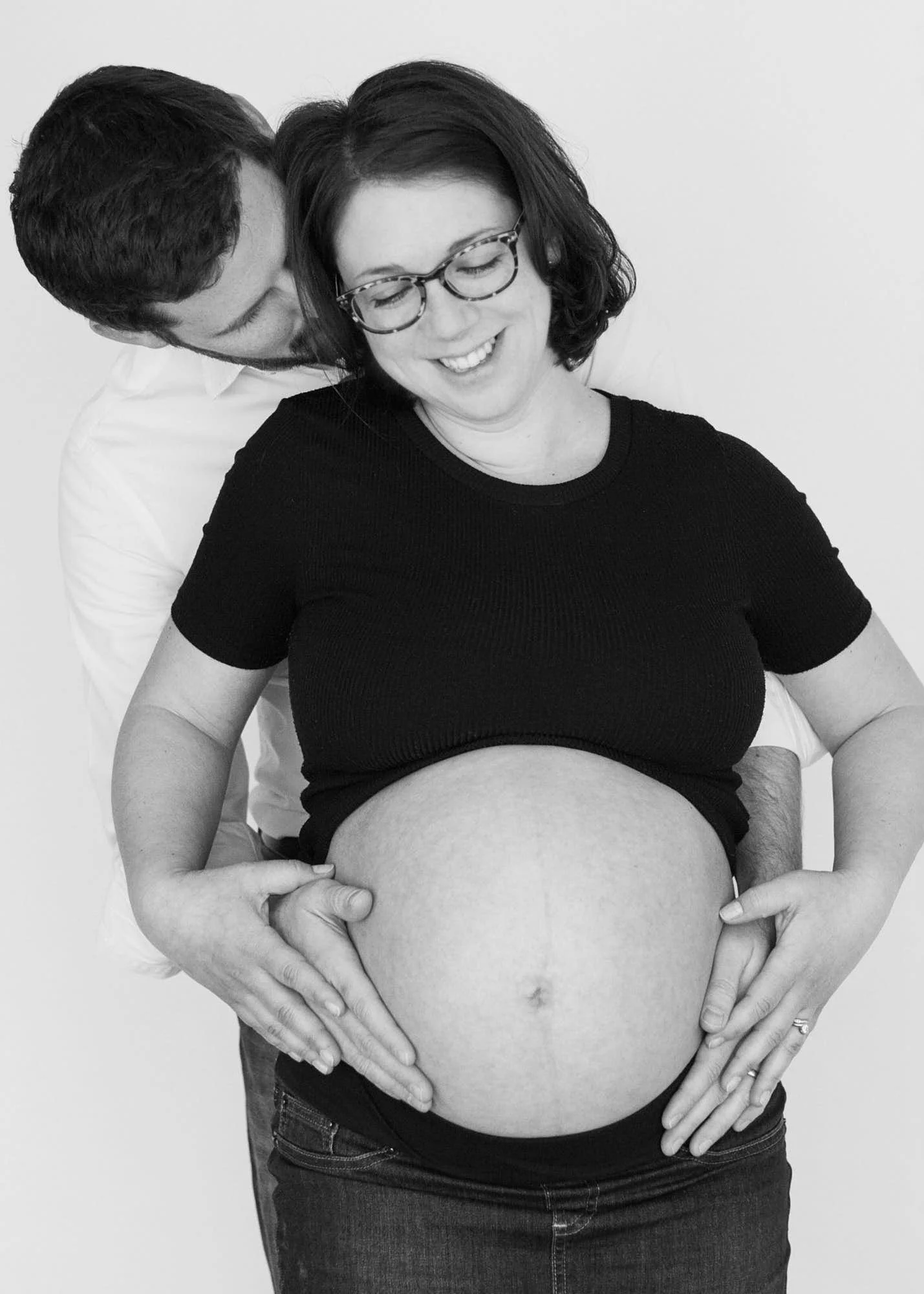 A pregnant woman smiling with glasses and dark hair, with her partner behind her, both with hands on her belly, in front of a plain white background.