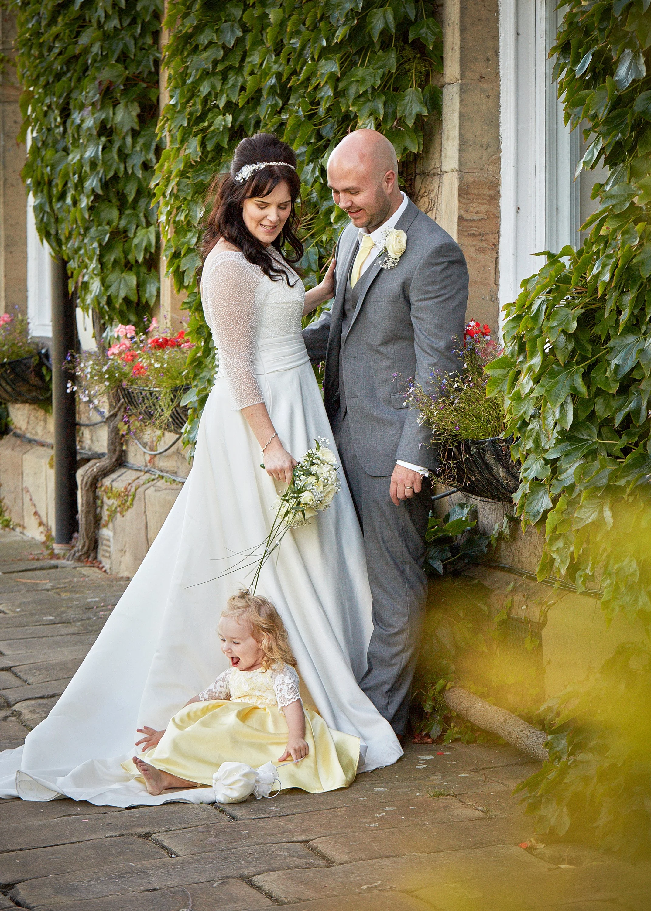 A bride and groom standing outside near a brick wall covered with green ivy, with two young girls sitting on the ground in front of them. The bride is wearing a white wedding dress and holding a small bouquet, and the groom is dressed in a gray suit with a white boutonniere. One girl in a yellow dress with lace sleeves is sitting with her legs extended, and another girl is partially visible with curly hair and a yellow dress.