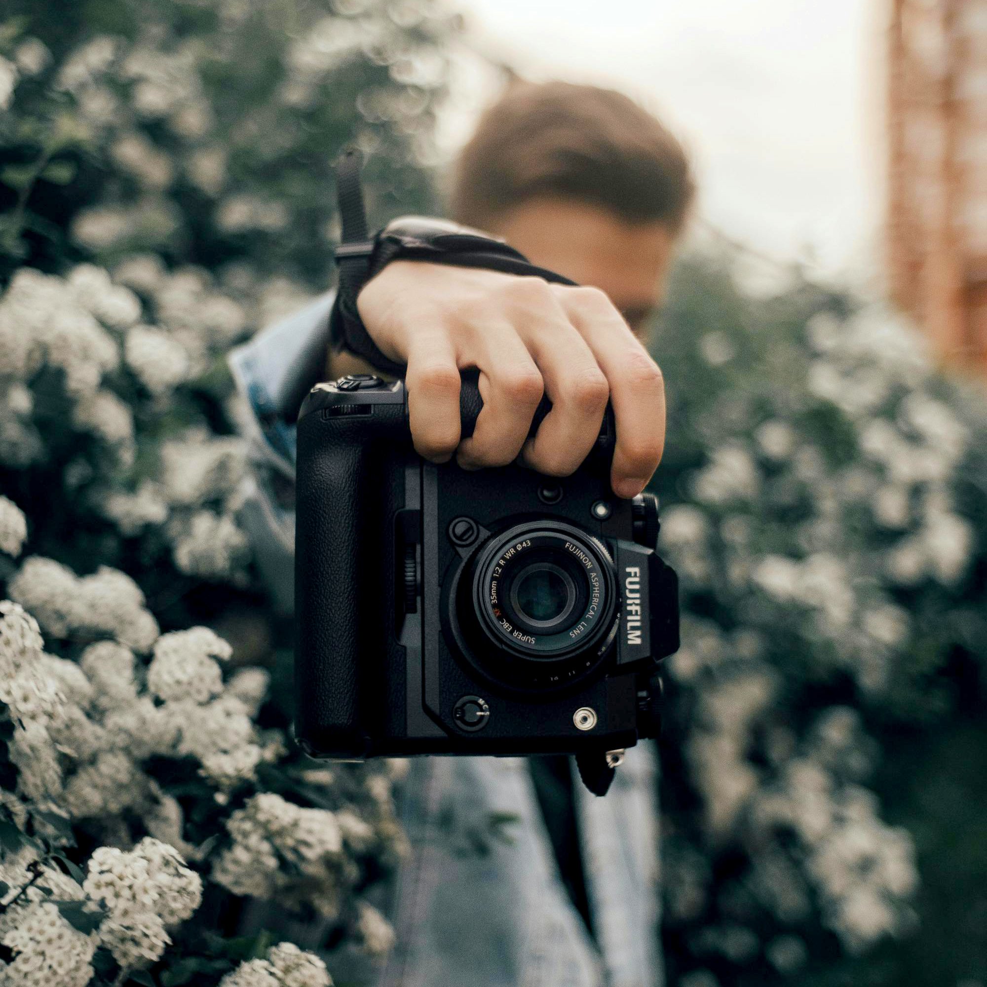 A person holding a camera close-up, blurred face, outdoor setting with trees and rocks.