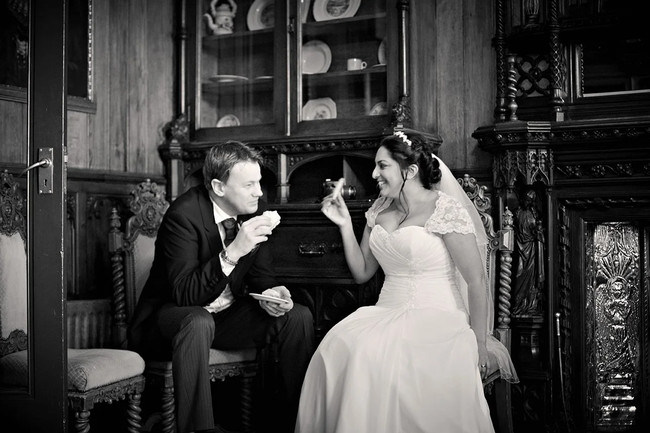 A bride and groom sitting indoors, smiling and engaging in conversation. The bride is wearing a wedding dress and a tiara, holding a slice of cake with her right hand. The groom is dressed in a suit, holding a plate and a fork. The setting appears to be a decorated room with dark wooden furniture and a cabinet filled with dishes in the background.