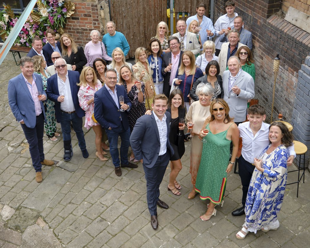 A group of around 30 people celebrating in a backyard, holding glasses of rosé, dressed in semi-formal attire, standing on stone paving with a brick wall and floral decorations in the background.