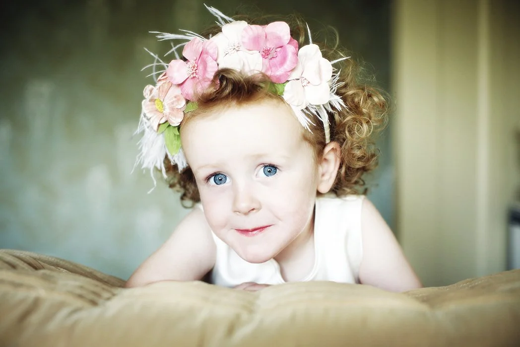 Young girl with curly red hair and blue eyes wearing a floral headband with pink and white flowers, resting her chin on a beige surface and looking at the camera.