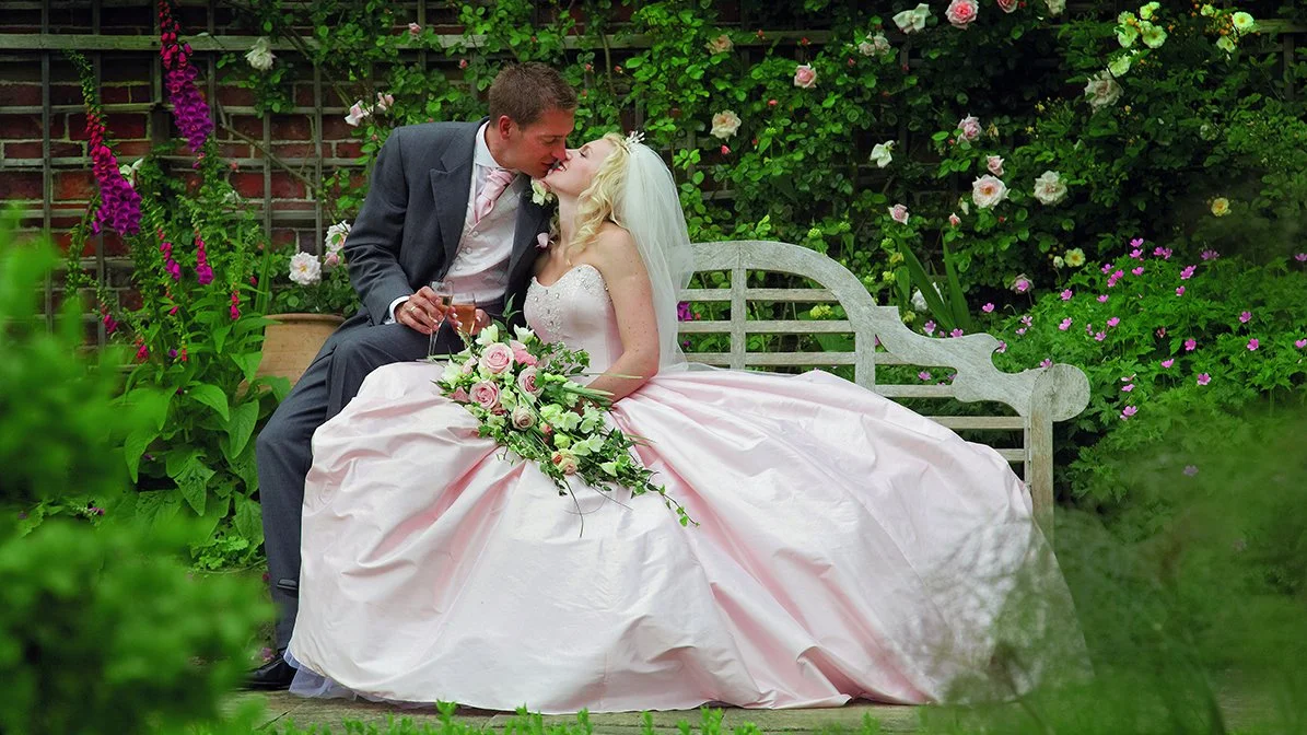 A newlywed couple shares a kiss in a lush garden, the bride in a pink wedding gown holding a bouquet of pink and white roses, and the groom in a dark suit with a glass of champagne.