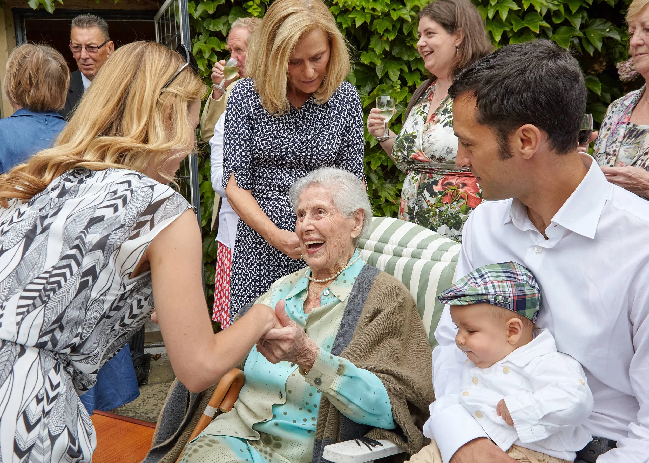 A group of people, including elderly and younger individuals, gathered outdoors around a seated elderly woman with white hair, who is smiling and holding hands with a woman in a patterned top. The scene shows a warm social gathering with drinks and l