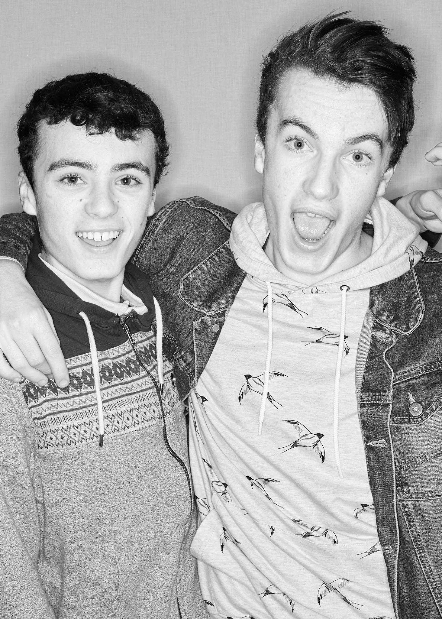 Two young men are smiling and posing with their arms around each other. The man on the right is making a surprised expression with his mouth open. They are dressed casually and are standing close together.