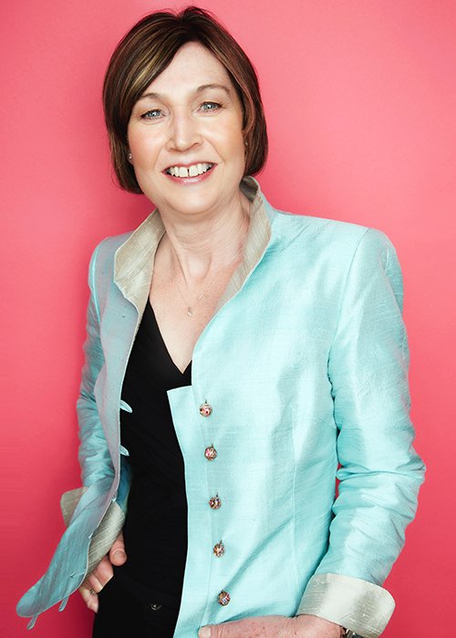 A woman with short brown hair and a friendly smile, wearing a light blue blazer with silver accents over a black top, standing against a pink background.