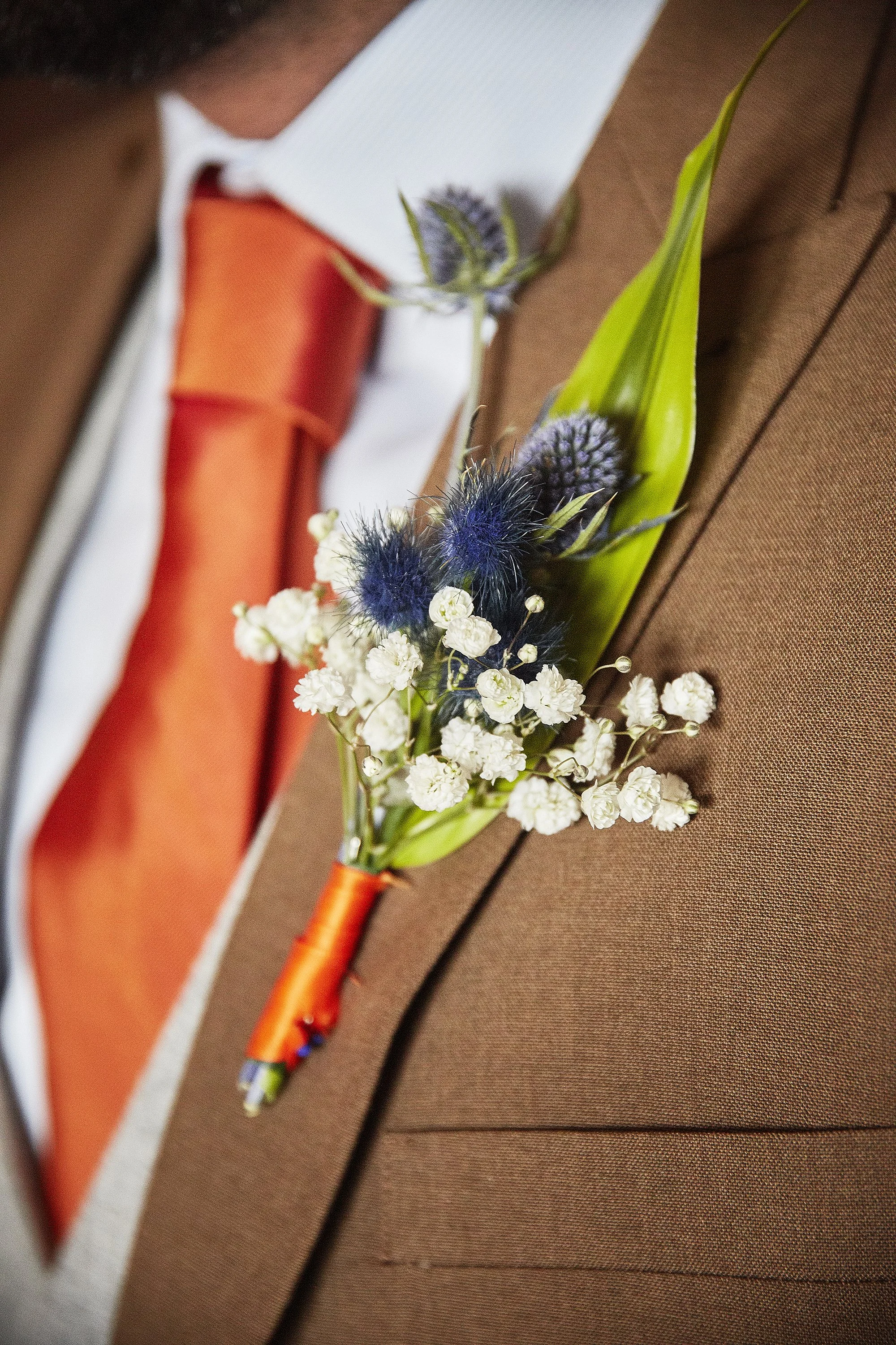 An orange boutonniere with white and purple flowers, dark blue thistles, and long green leaves, attached to a brown suit jacket.