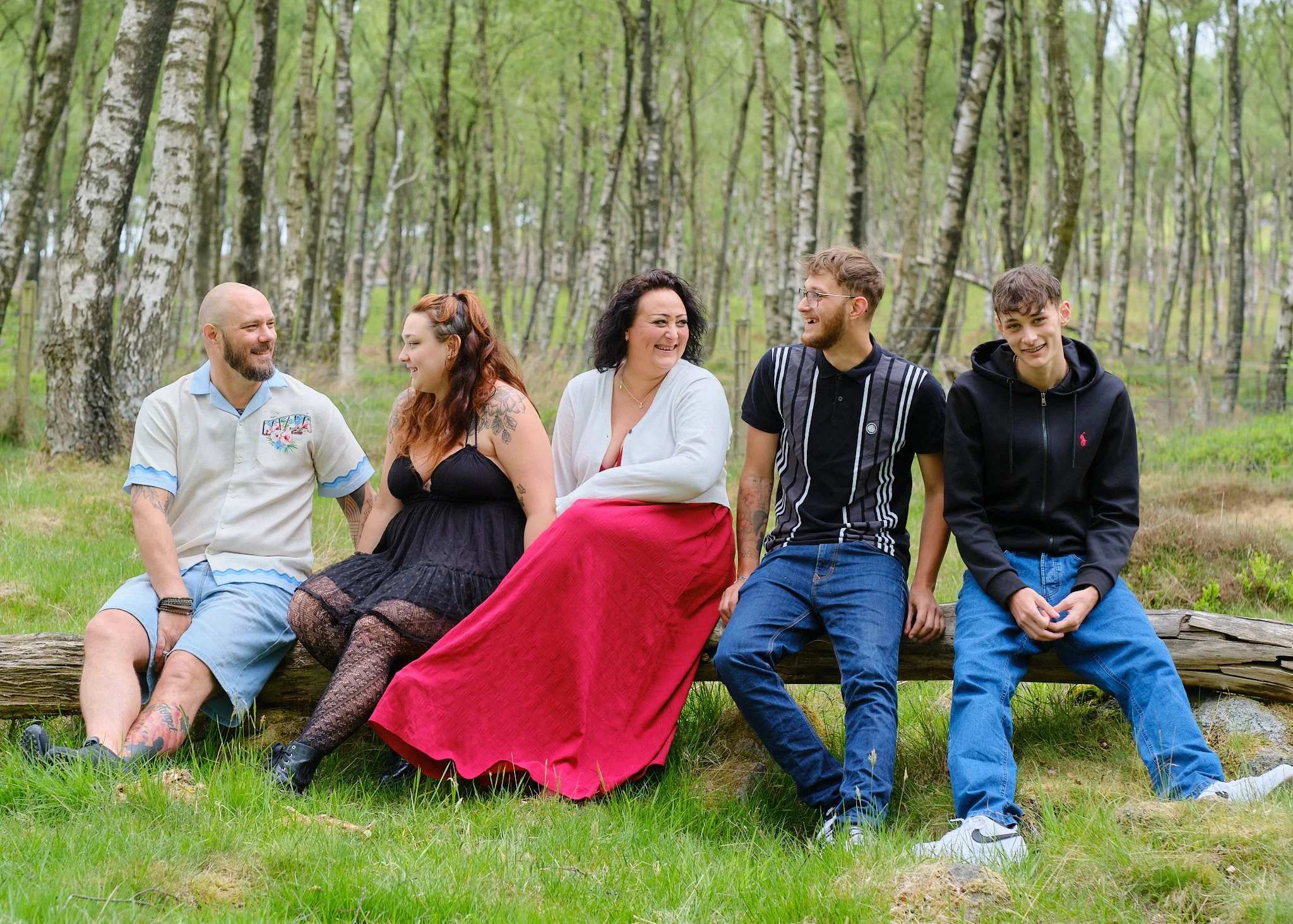 Six people sitting on a log in a green forest, smiling and talking to each other.