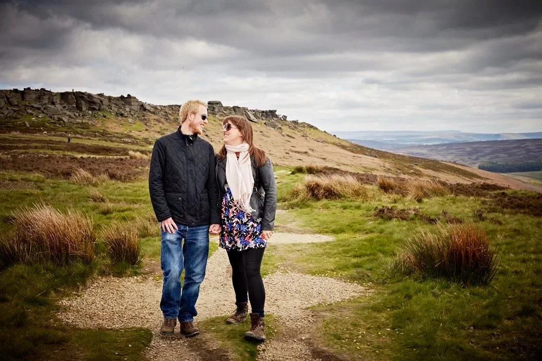 A couple walking and holding hands on a dirt path in a hilly landscape with grass, shrubs, and rocks, under a cloudy sky.
