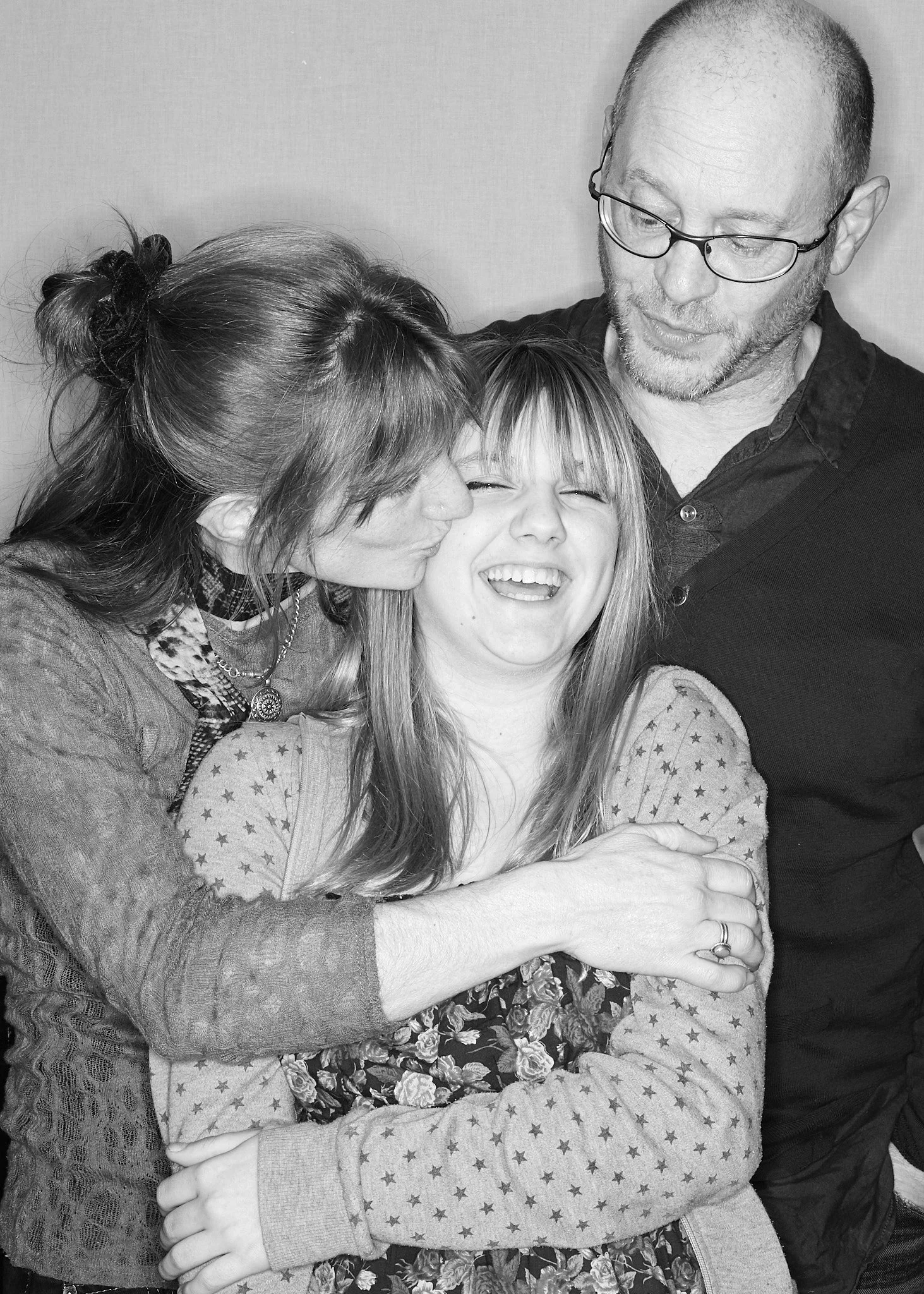 A man with glasses and a beard faces downward and smiles, a woman kisses his cheek, and a girl with blonde hair and star-patterned shirt smiles with her eyes closed as they hug.