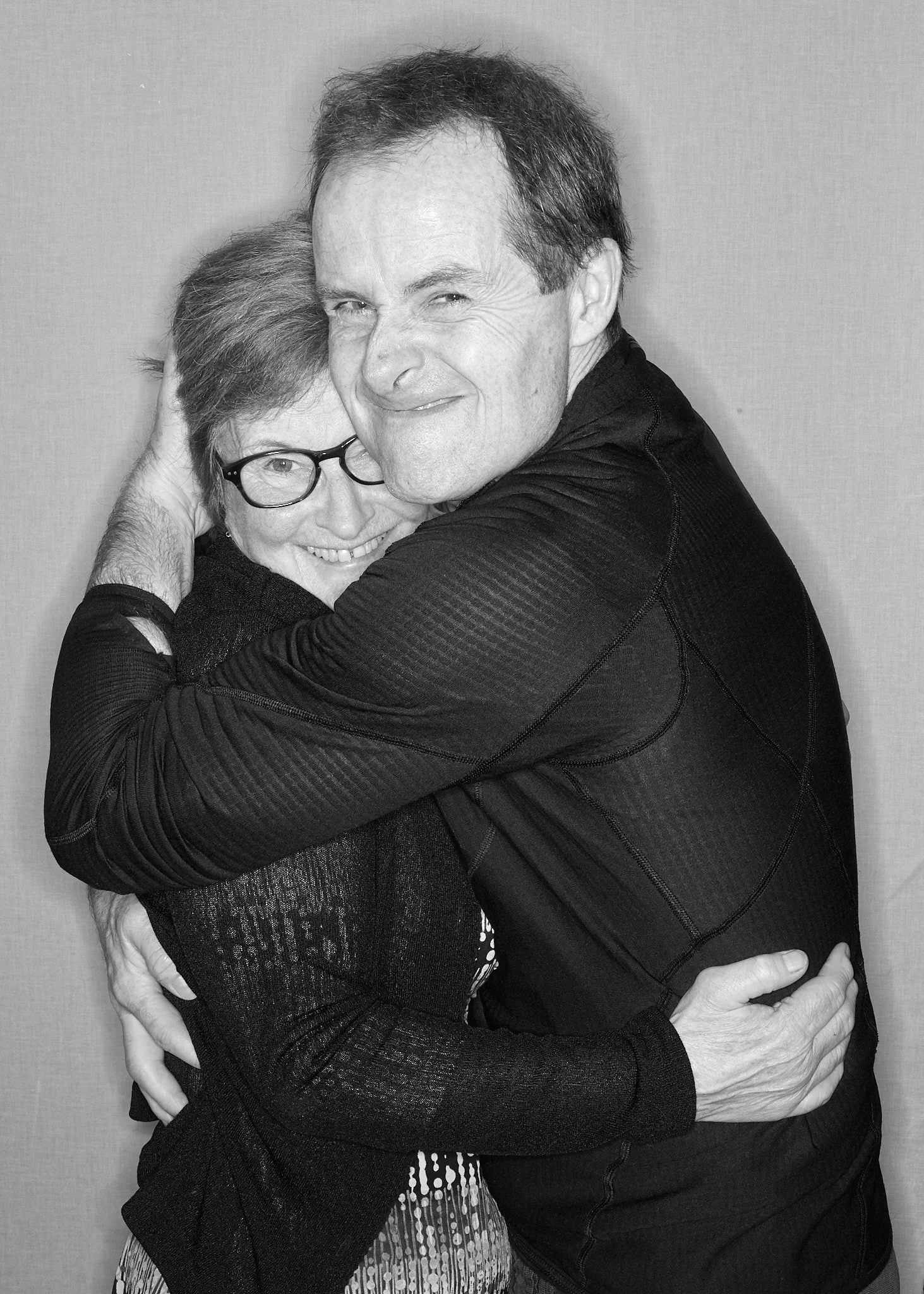 A black and white photo of two people hugging warmly. The person on the left is an older woman with glasses and short hair, smiling happily. The person on the right is a middle-aged man with short hair, smiling gently. They are standing close togethe