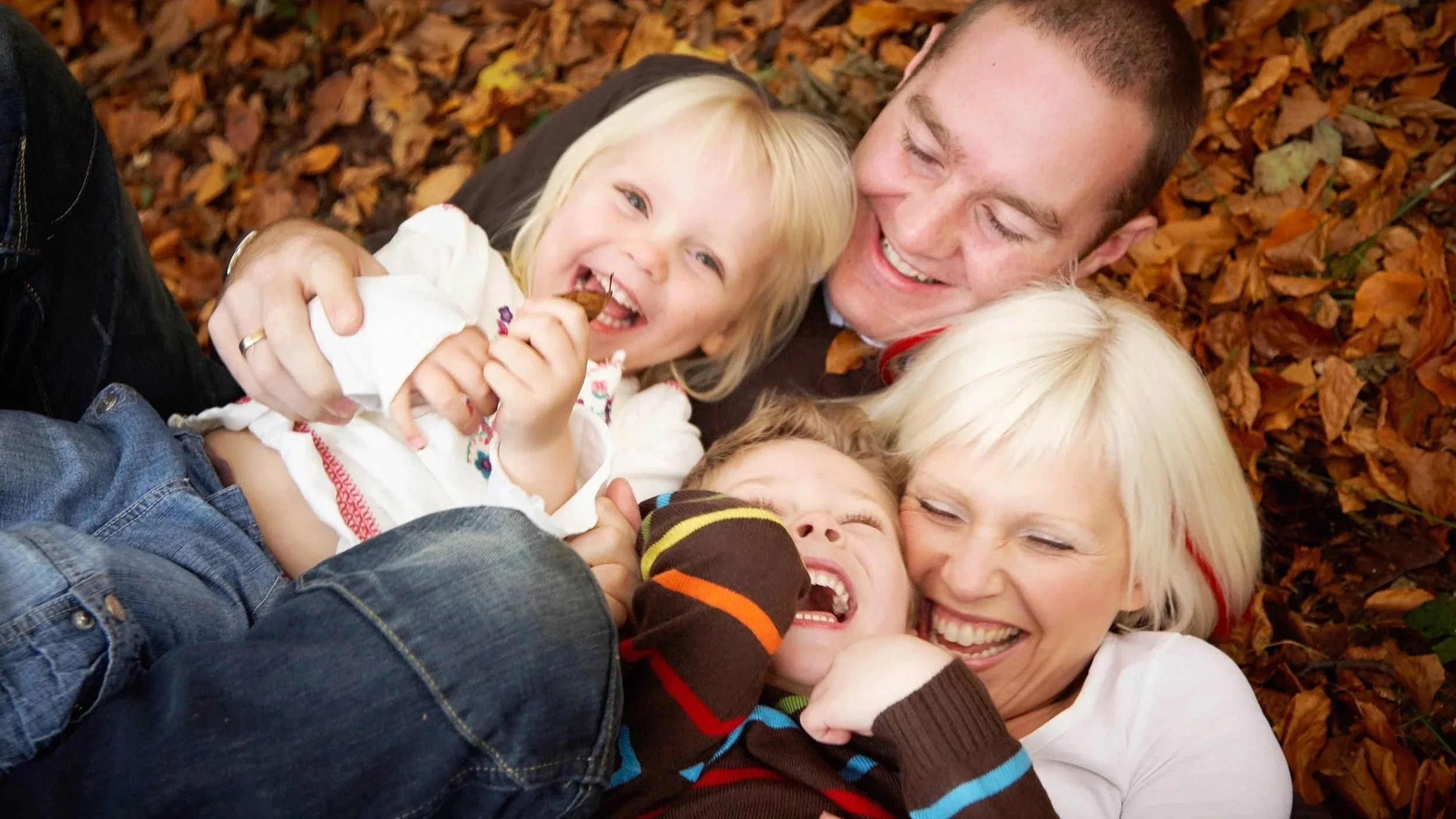 A group of children and adults lying on a bed of autumn leaves, smiling and laughing together.