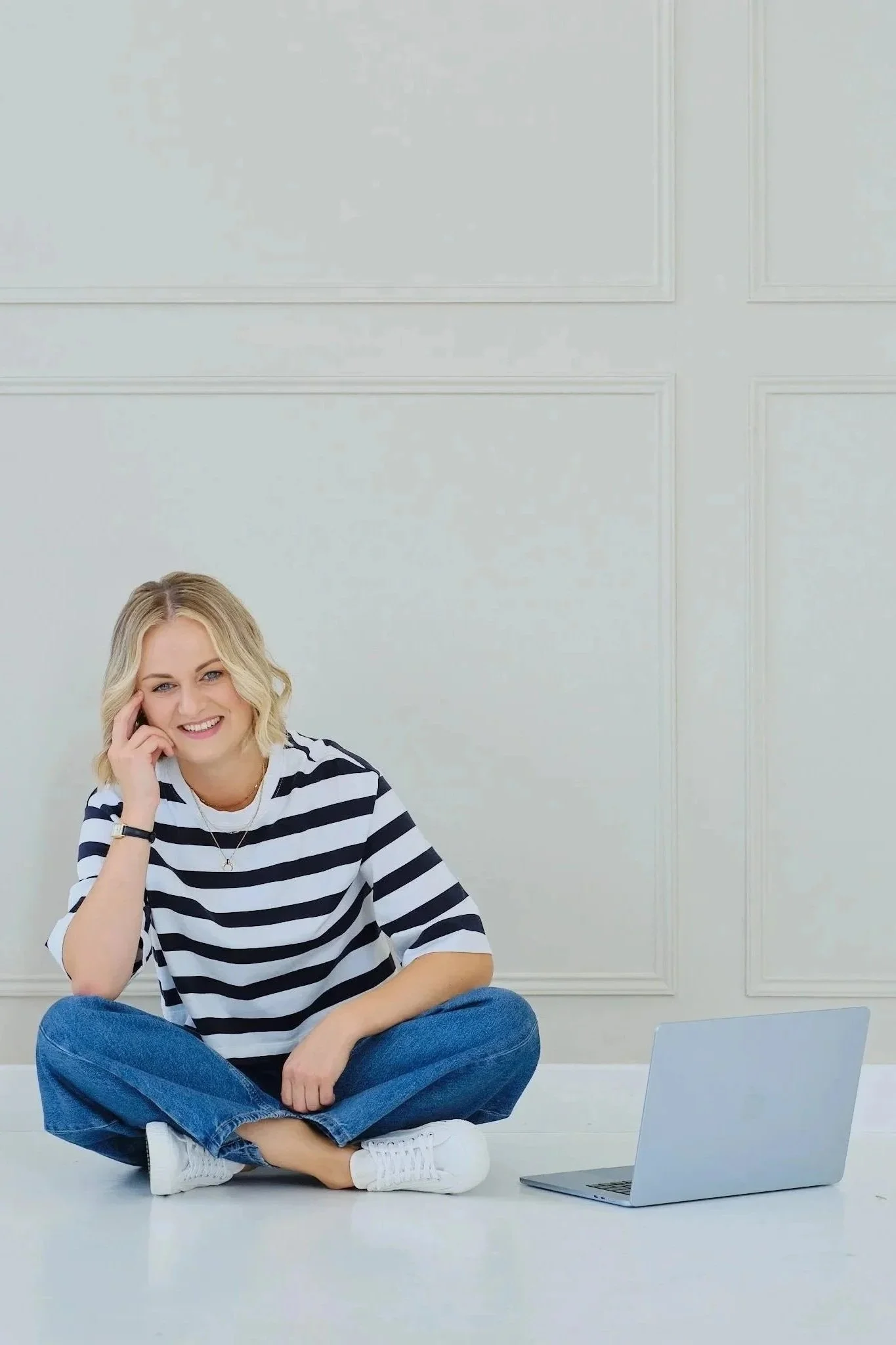 A woman sitting cross-legged on the floor with a laptop open in front of her, smiling, wearing a striped shirt and blue jeans, in a minimalistic room with white walls.