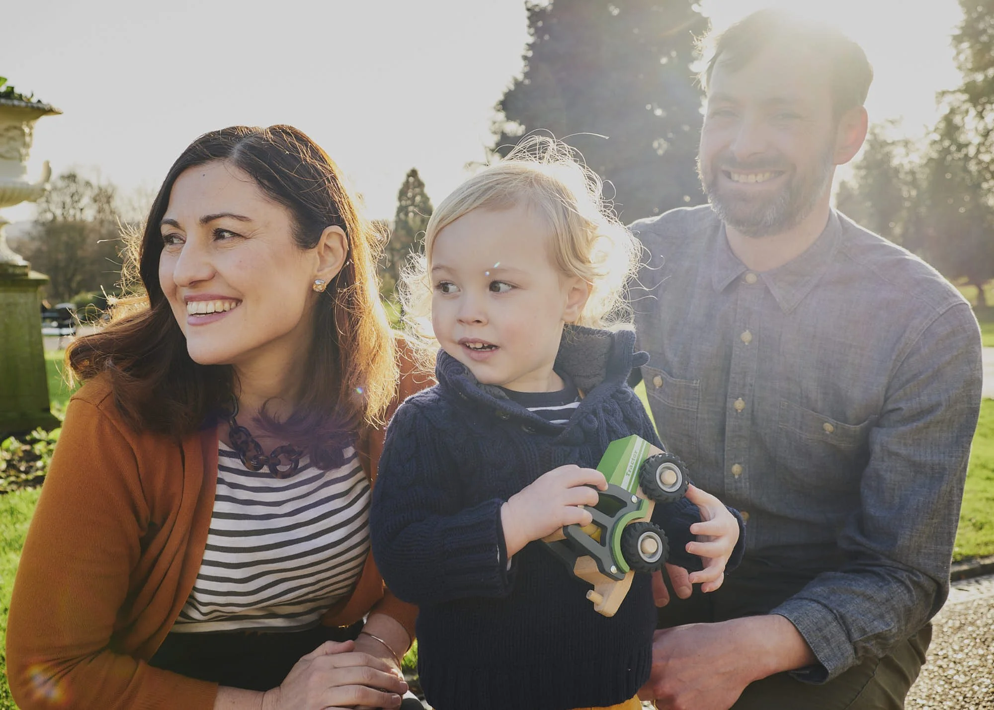 A smiling family of three, including a woman with long brown hair, a young boy with blond hair holding a toy car, and a man with short brown hair, outside on a sunny day in a park with trees in the background.
