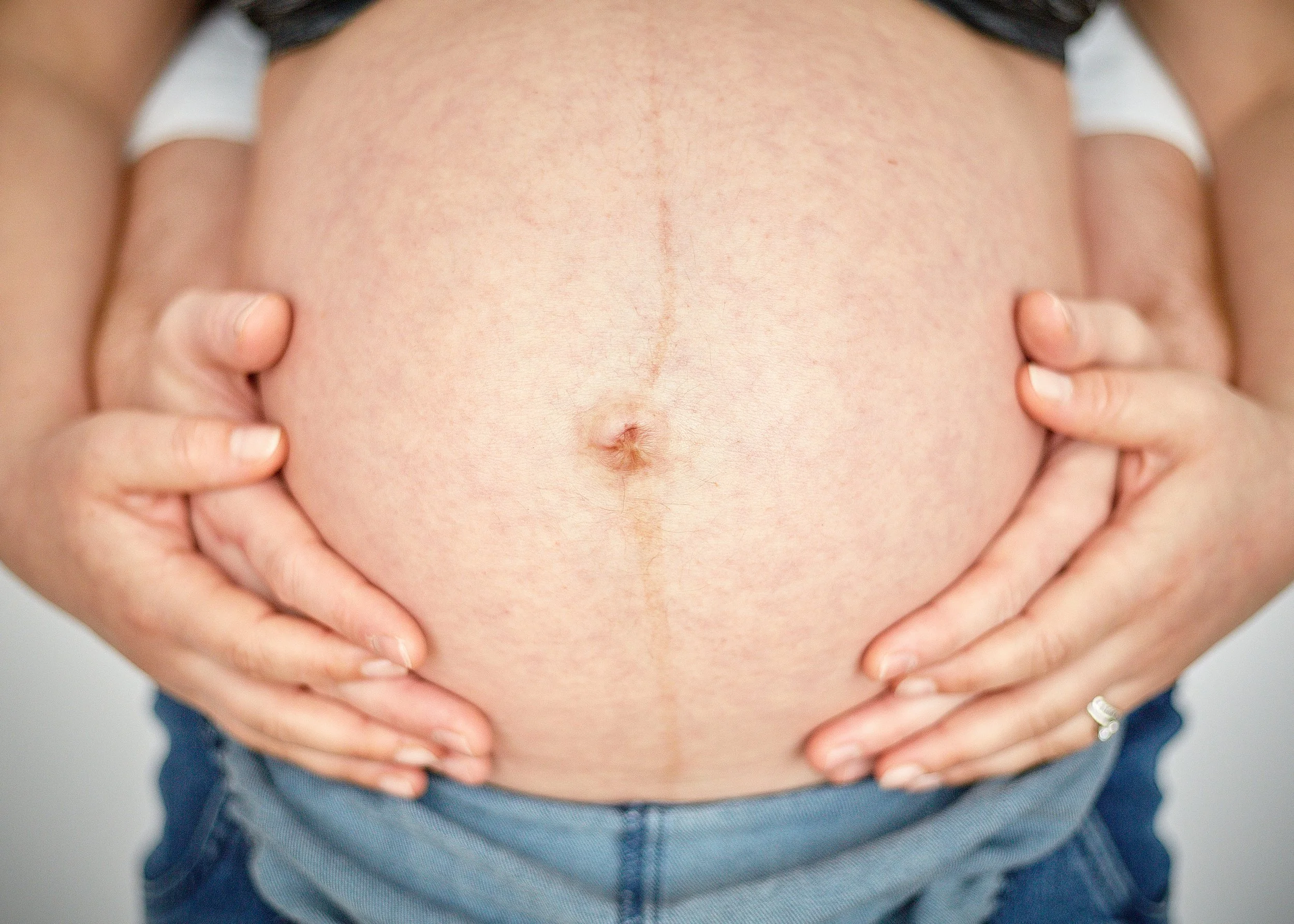 Person holding their pregnant belly with both hands, wearing a dark shirt and blue jeans.