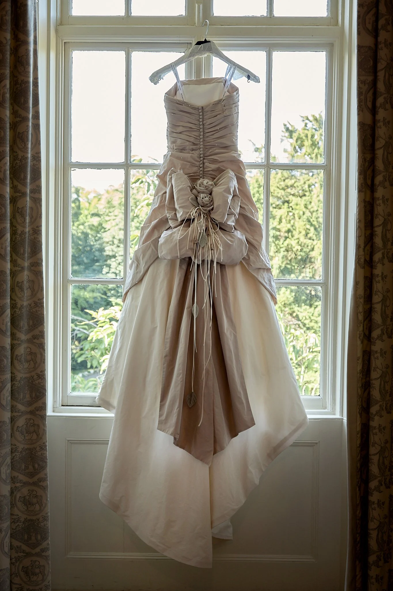 A wedding dress hanging on a hanger in front of a window with greenery outside.