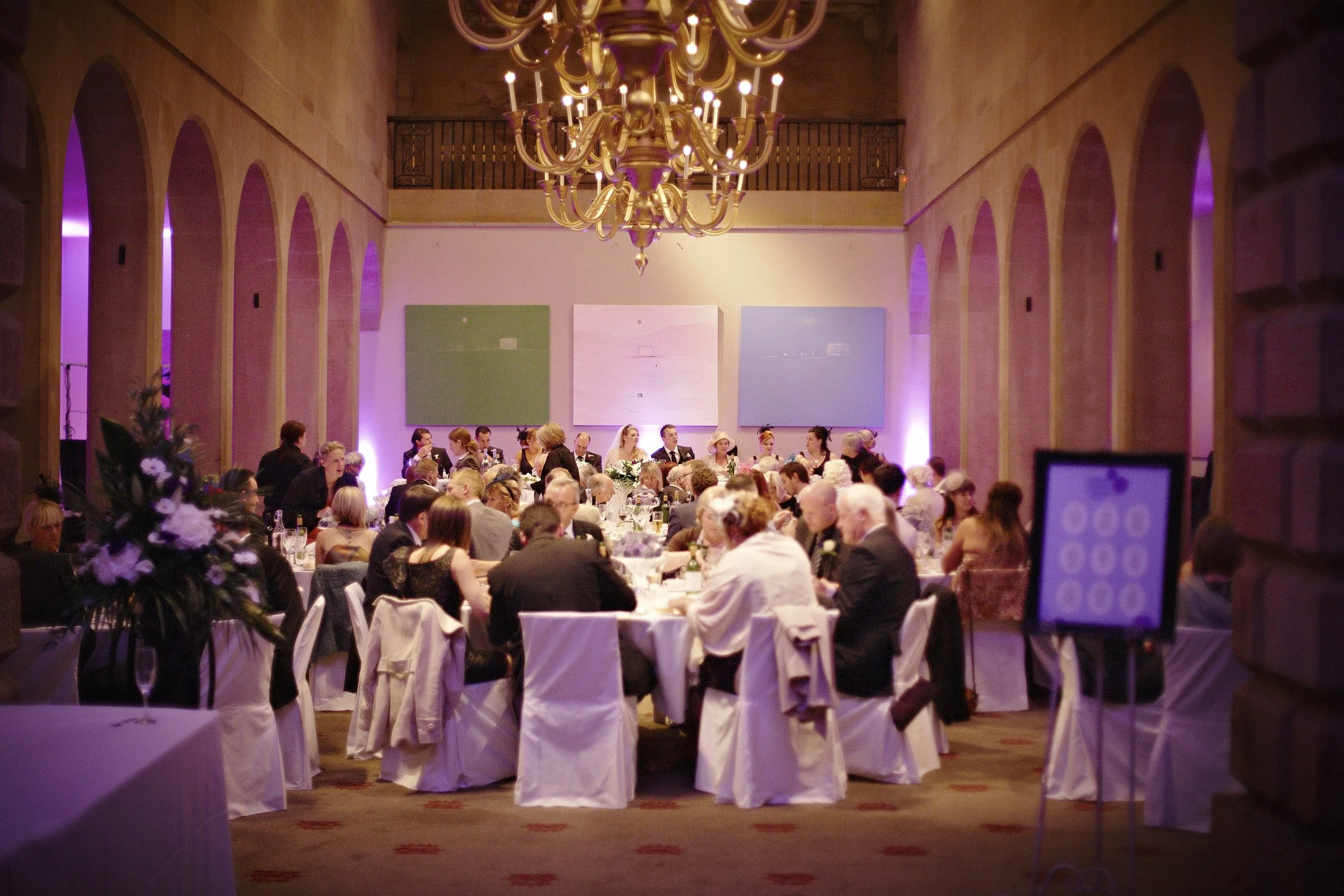 A large banquet hall with many round tables covered in white tablecloths, set with  elegant place settings, and surrounded by people dressed in formal attire, attending a wedding or celebratory event. The ceiling has large arches with purple lighting