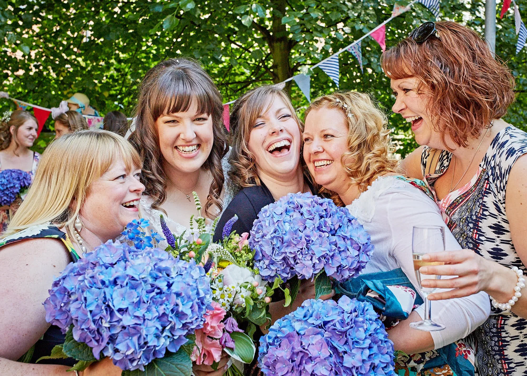 A group of women and girls smiling and hugging outdoors during a celebration, with colorful bunting and greenery in the background, and blue hydrangea flowers in the foreground.