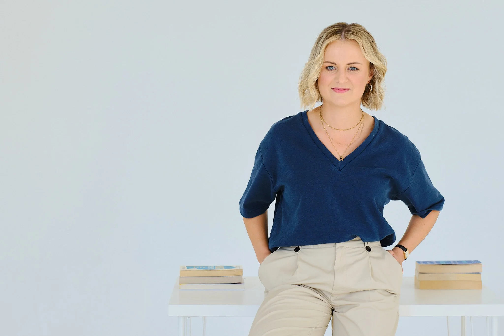 A woman with blonde hair wearing a blue top and beige pants, sitting on a white table with stacks of books on either side, against a plain white background.